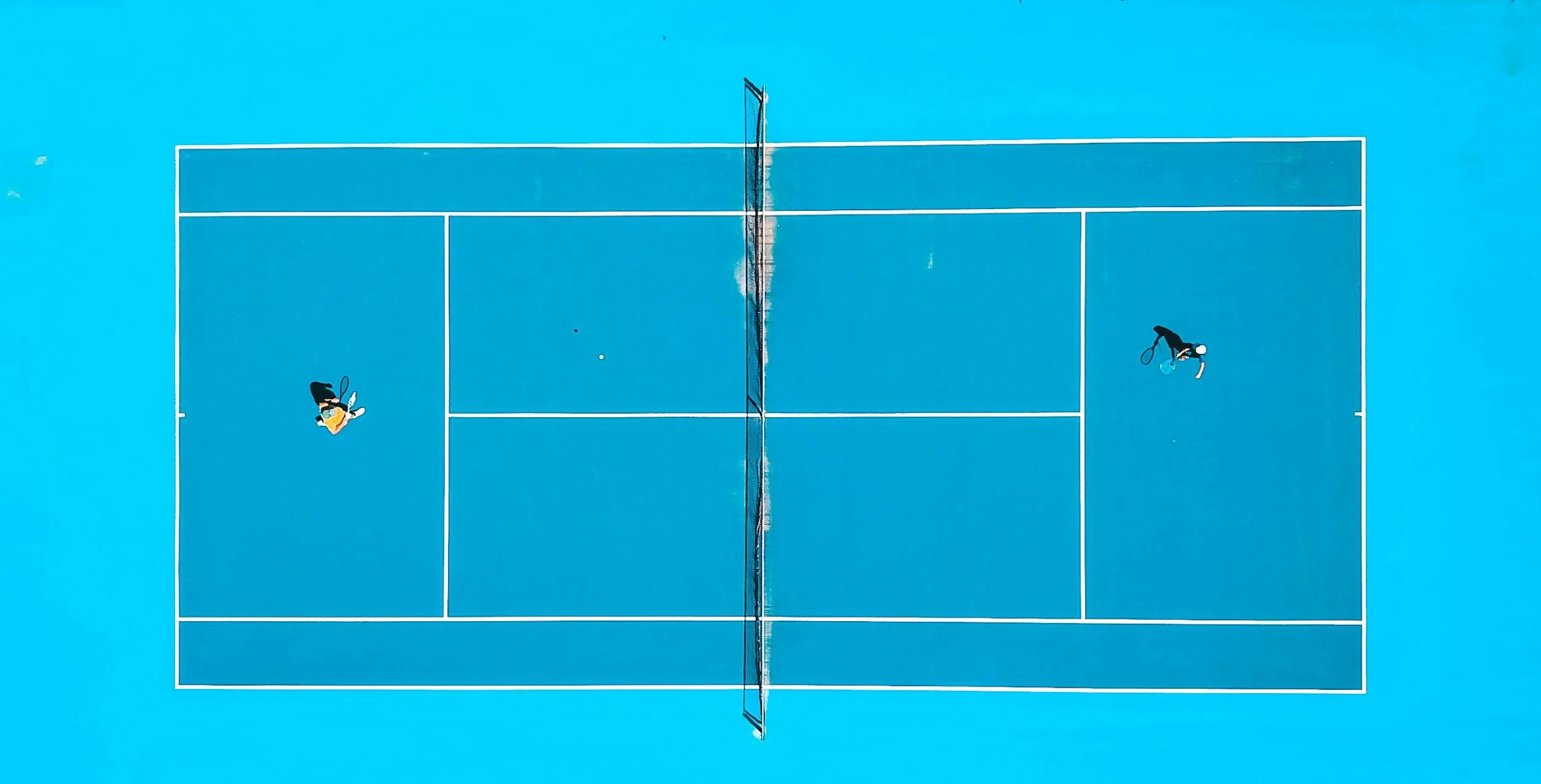 Top-down view of a blue tennis court with two players preparing to serve, separated by a net.
