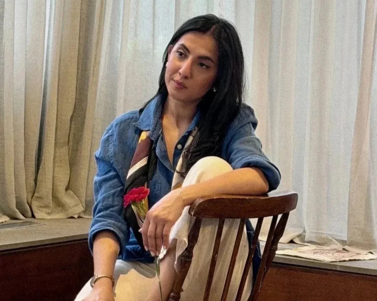 A woman sitting on a wooden chair, wearing a blue shirt with rolled-up sleeves, holding a red flower in her hand, with a pensive expression, in front of cream-colored curtains.