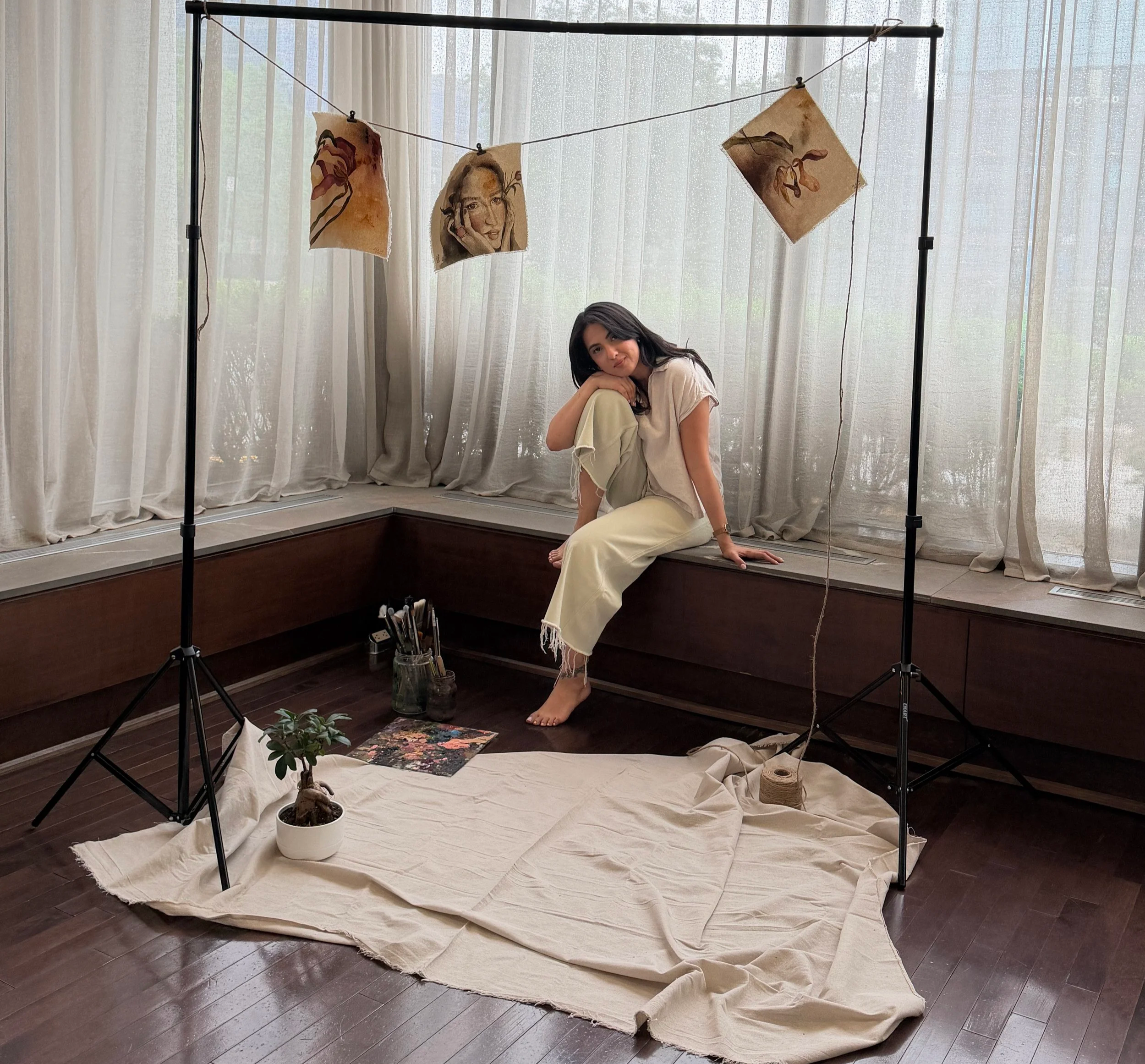 Art installation with three watercolor portraits hanging on a wire, a woman sitting on a wooden ledge in front of sheer curtains, houseplants, and art supplies on a white cloth on the floor.