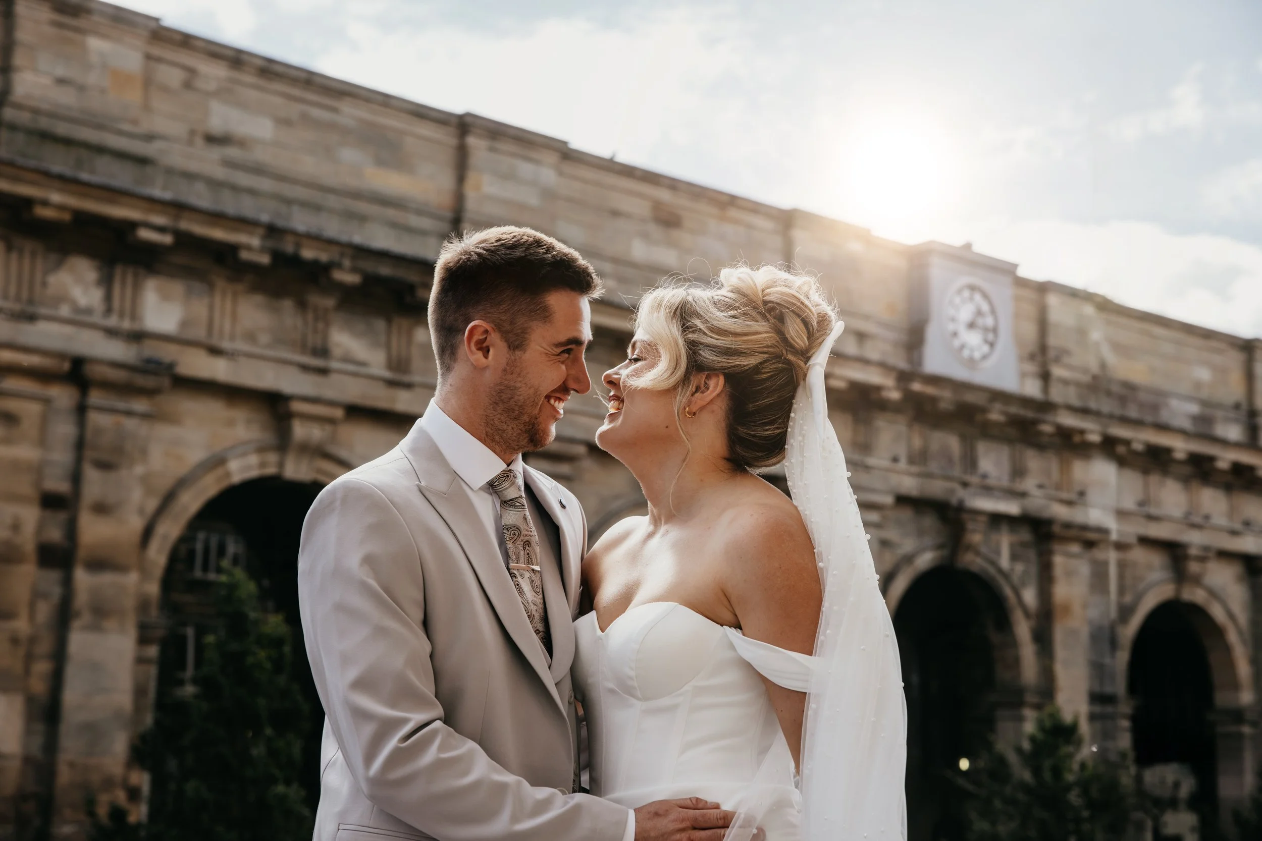 A newlywed couple, a man in a white tuxedo and a woman in a white wedding dress, smiling and staring into each other's eyes outdoors in front of a historic stone building with a clock, during what appears to be late afternoon or early evening.