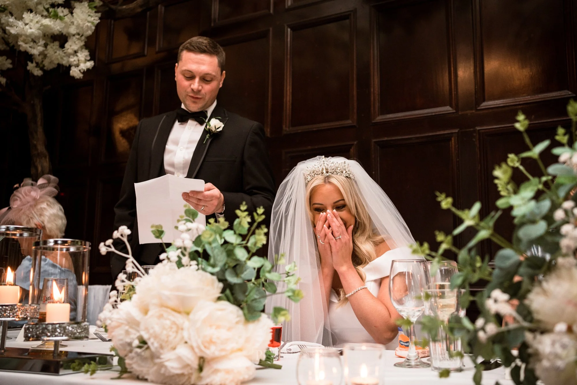 Bride covering her mouth while seated at a wedding reception table, with a man standing beside her reading from a paper, decorated with white flowers and candles, in a wood-paneled room.