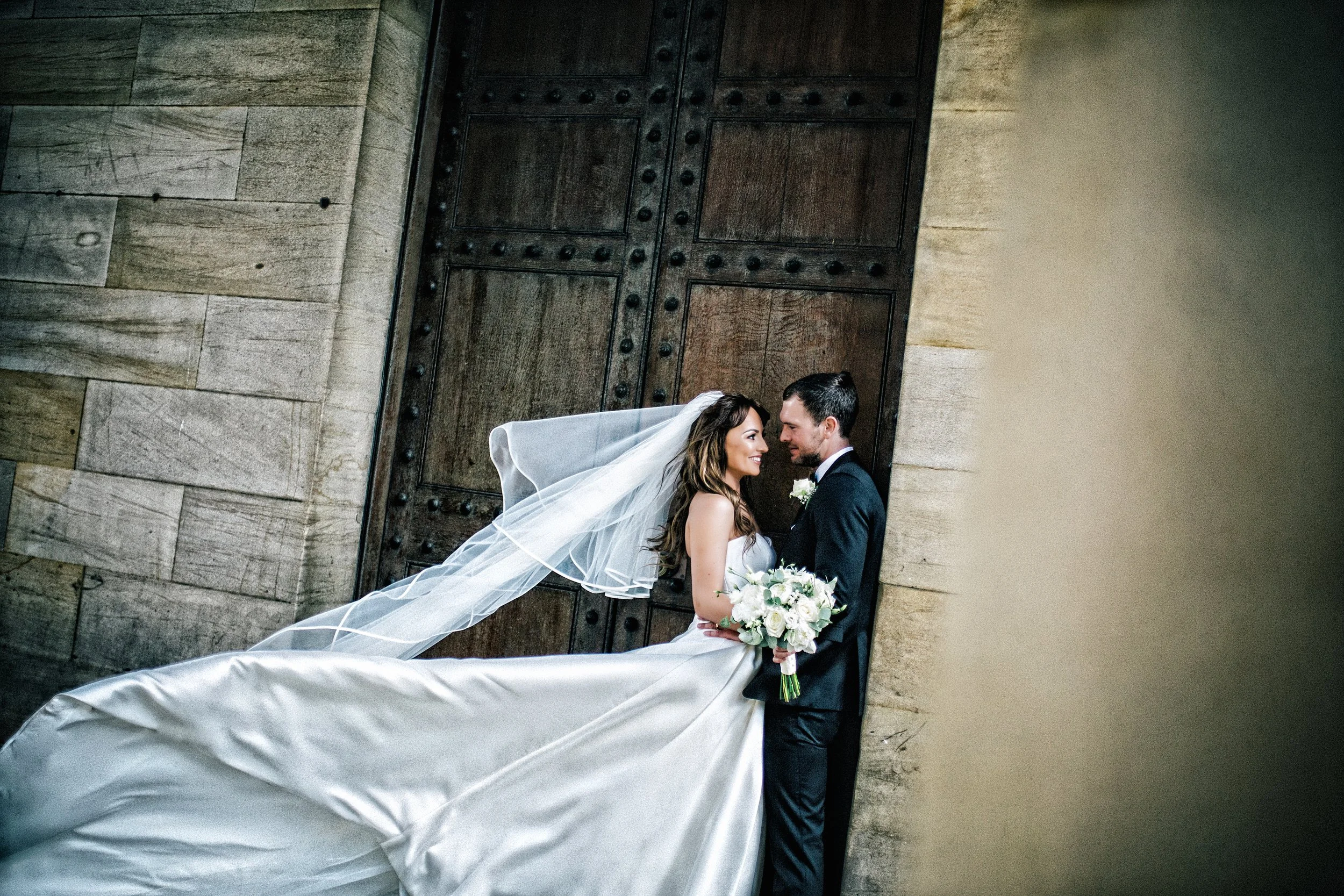 A bride and groom stand close to each other in front of a large wooden door, smiling and gazing into each other's eyes. The bride is holding a bouquet of white flowers, and she is wearing a white wedding dress with a long veil. The groom is dressed i