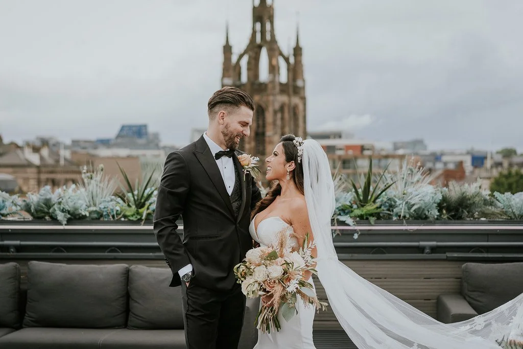 A bride and groom on a rooftop with city buildings in the background, smiling at each other. The bride is holding a bouquet and wearing a white wedding dress with a veil, and the groom is in a black tuxedo with a boutonniere.