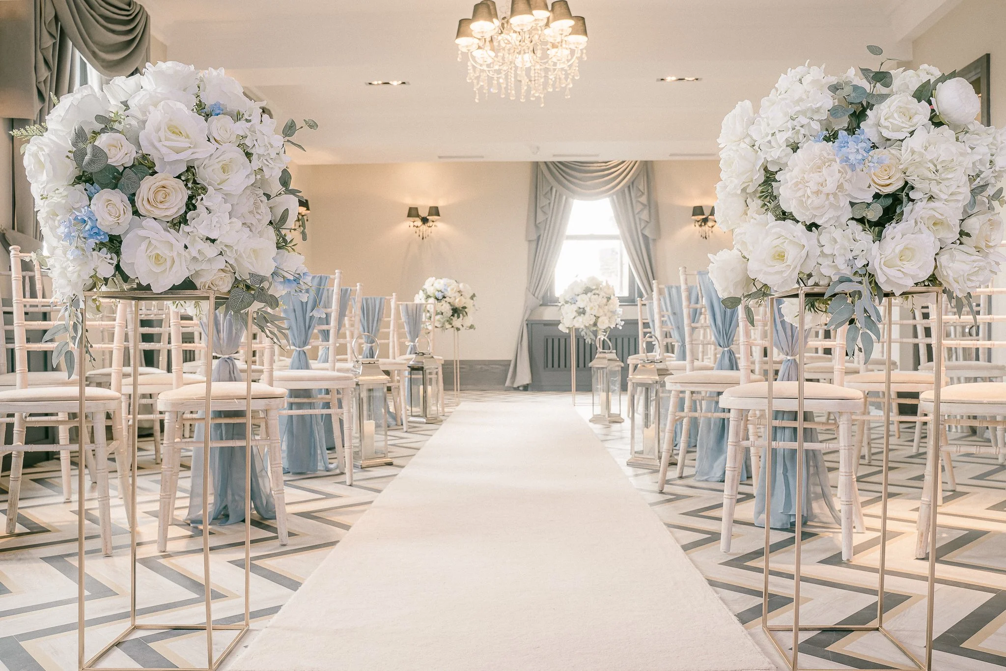 Elegant wedding ceremony setup with white chairs, tall floral arrangements of white roses and hydrangeas, and a white aisle runner in a decorated hall with chandelier and draped curtains.