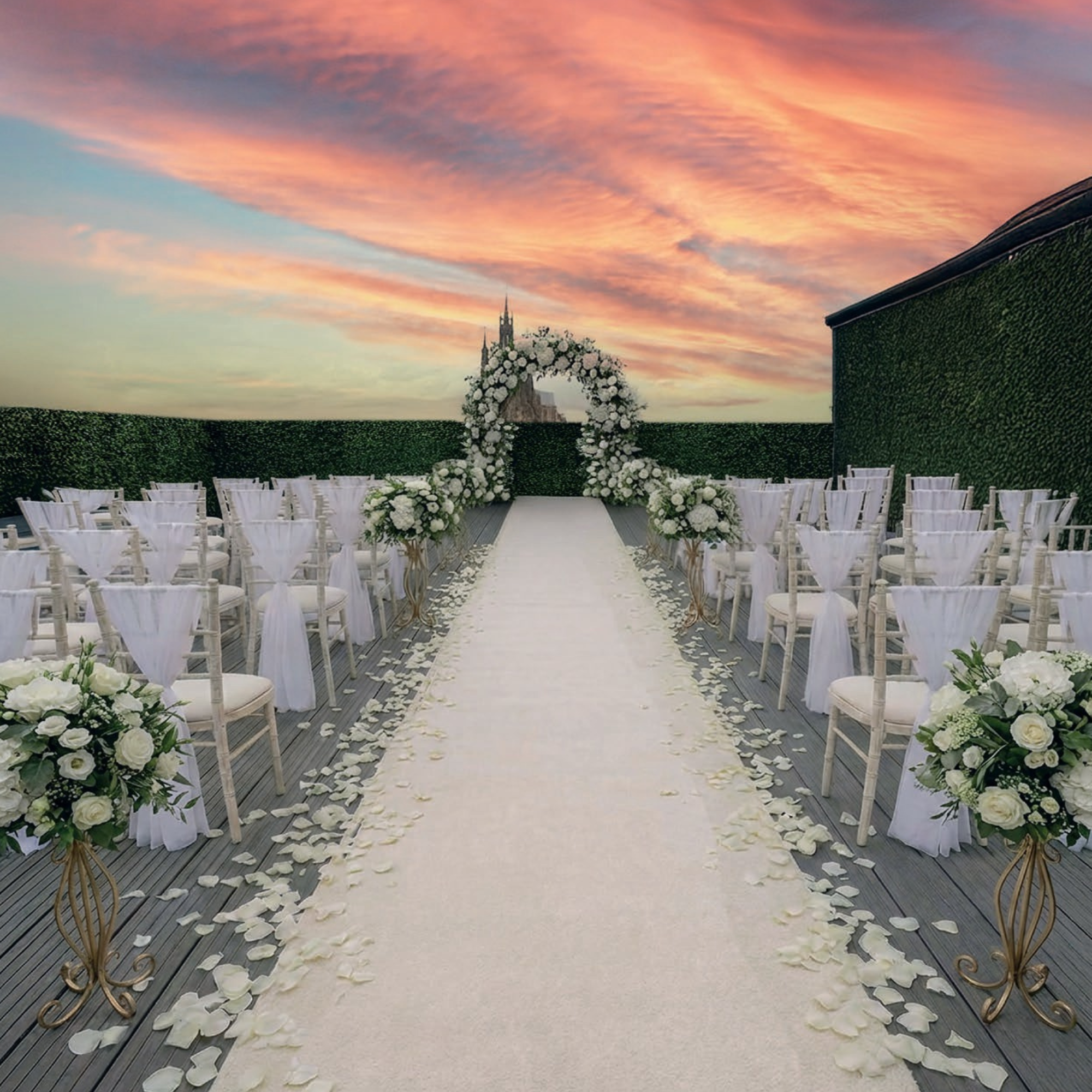 Wedding ceremony setup with white chairs, floral decorations, and an arch, set on a rooftop with a sunset sky.