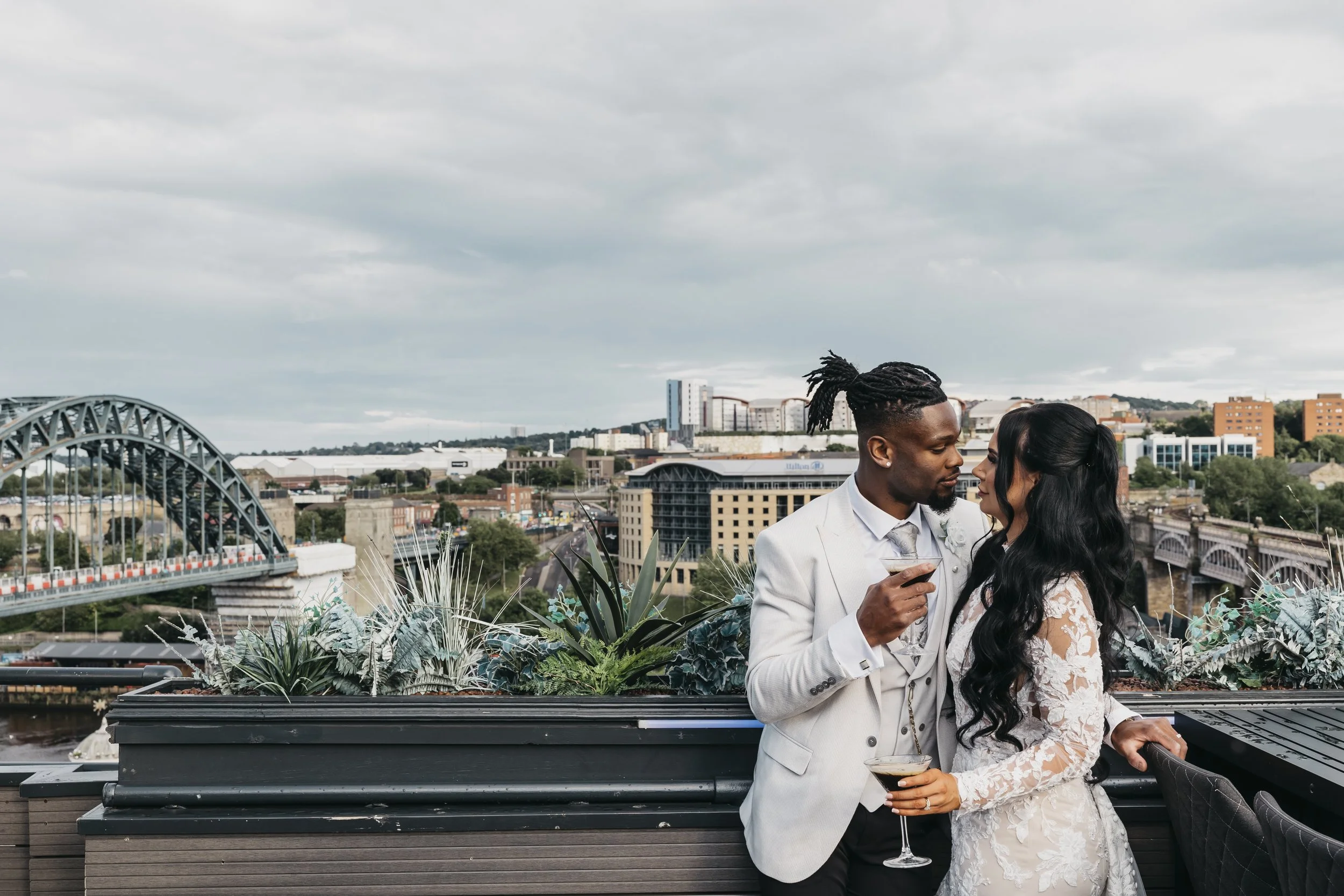 A romantic couple dressed in wedding attire sharing a moment on a rooftop with cityscape, bridge, and buildings in the background.