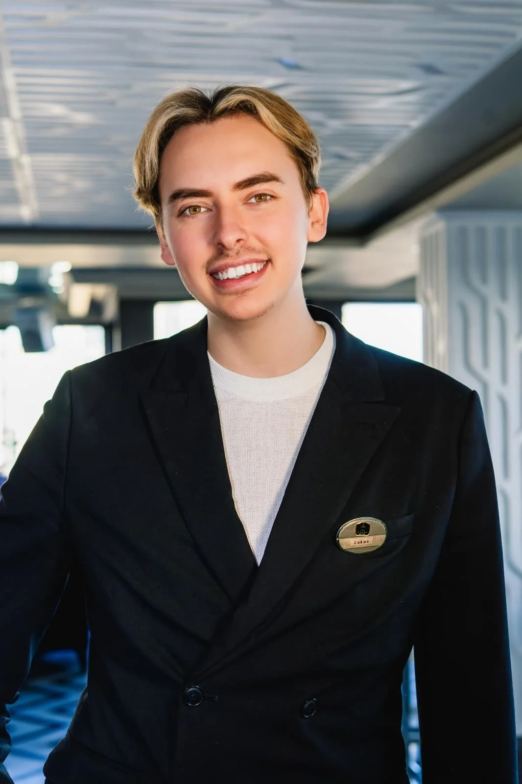 Young man with blonde hair and a light complexion smiling in a professional setting, wearing a black suit jacket with a name tag.