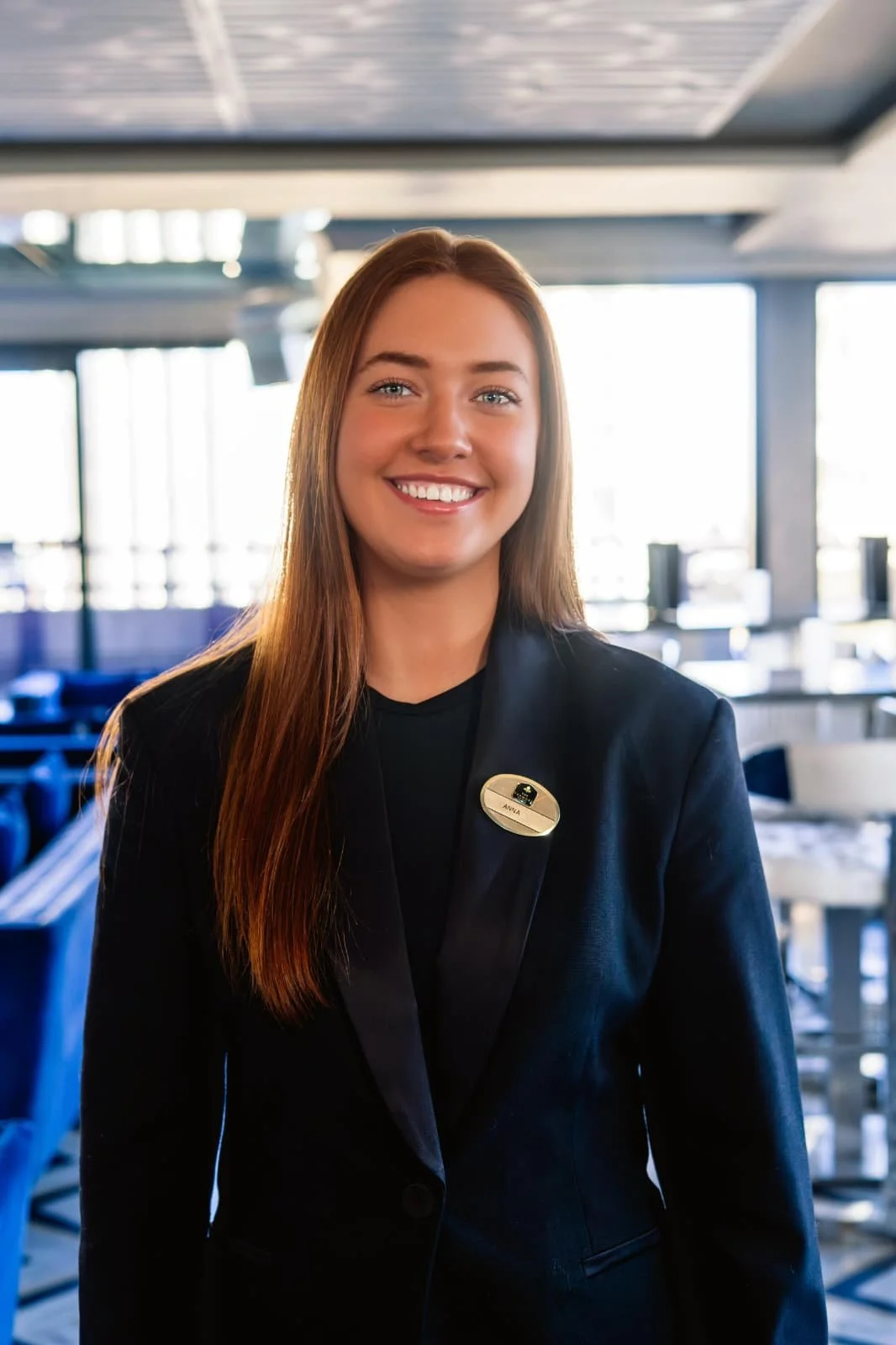 A smiling young woman with long red hair, dressed in a black blazer with a name tag, standing indoors in a modern restaurant or café with large windows and bright natural light.