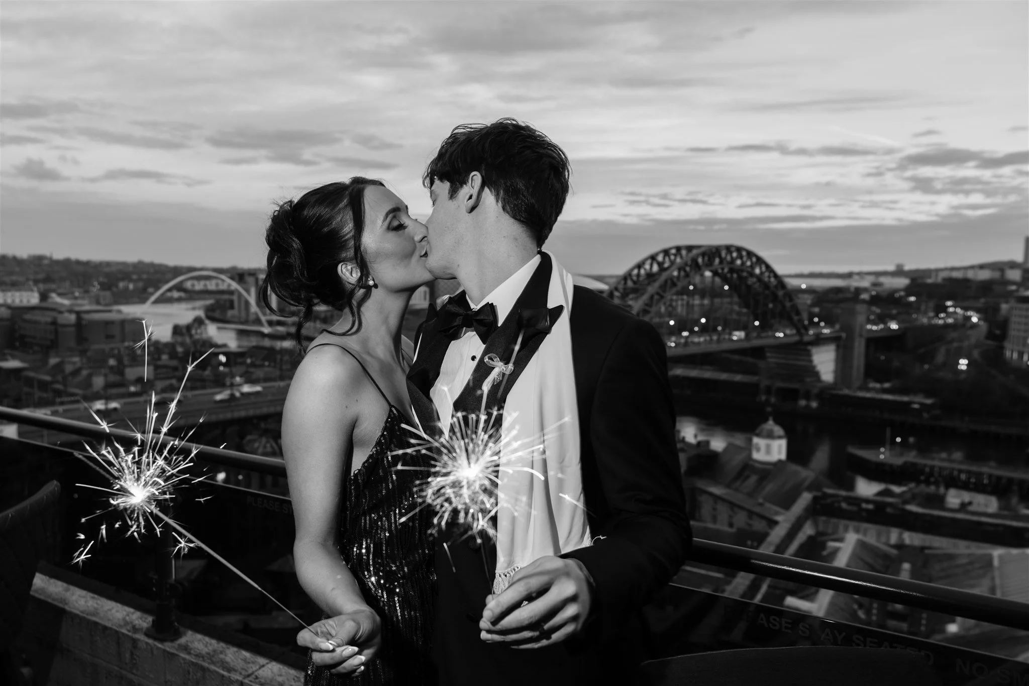 A newlywed couple in formal attire sharing a kiss on a rooftop with sparklers, city skyline, and bridge in background, in black and white.