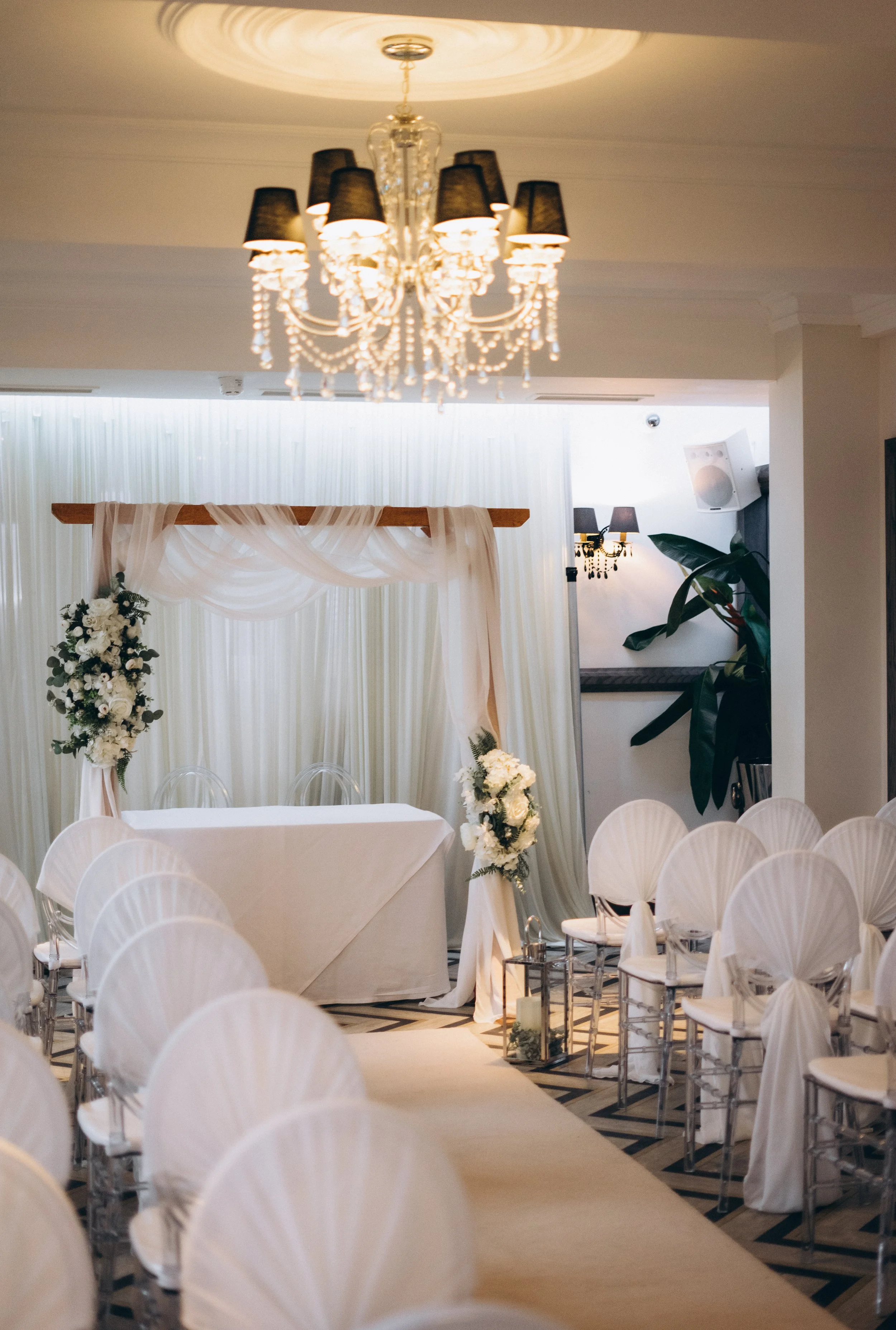 Elegant wedding venue with a chandelier, decorated table with white tablecloth, white chairs, and floral arrangements, featuring draped fabric backdrop and soft lighting.