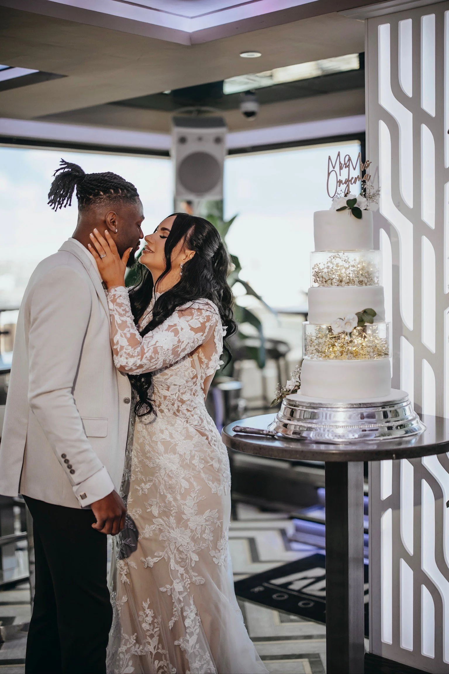 A newlywed couple, a man in a light-colored suit and a woman in a white lace wedding dress, share a kiss at their wedding reception next to a four-tiered wedding cake decorated with white flowers and greenery.