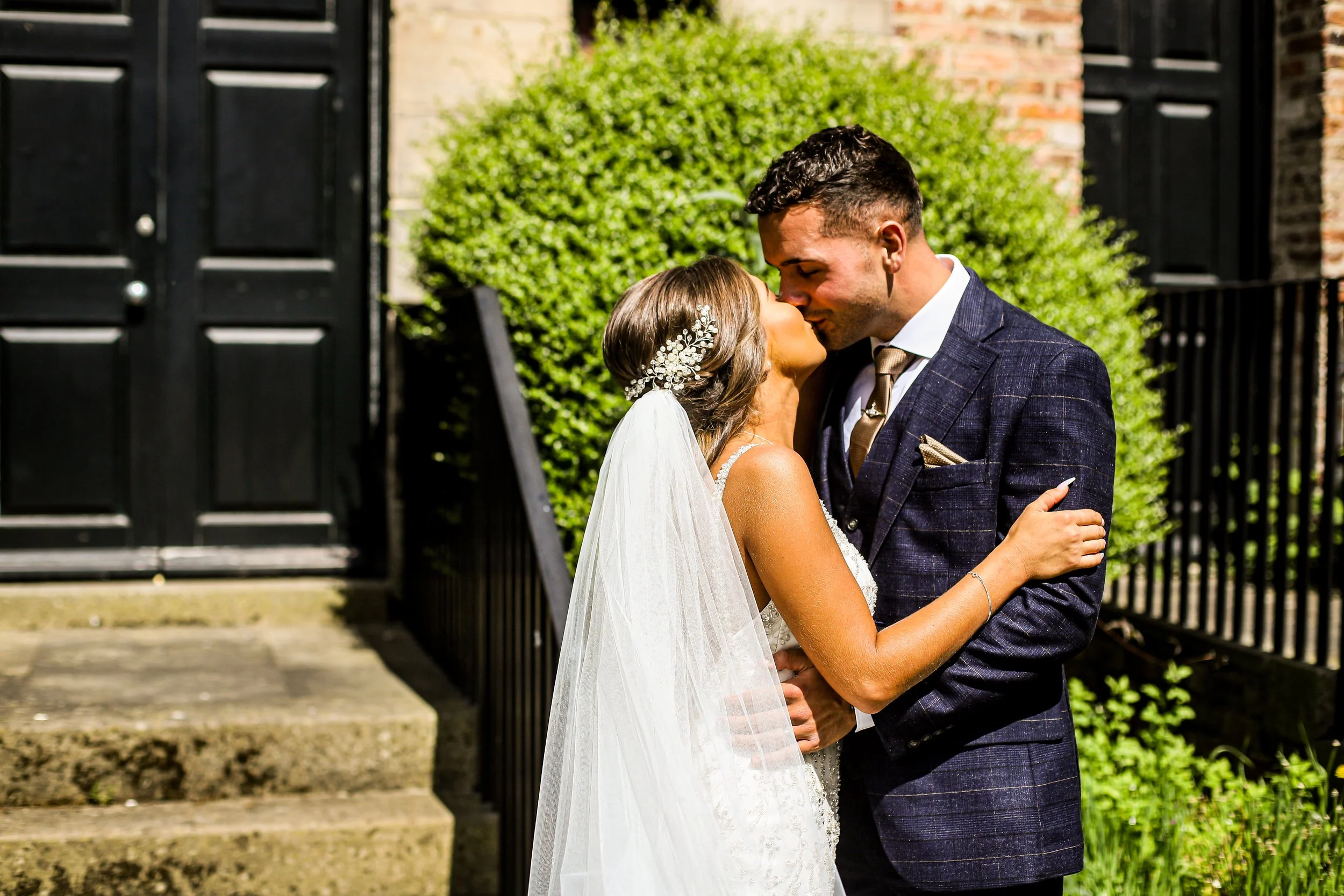 A bride and groom sharing a kiss outdoors during their wedding. The bride is wearing a white dress with a veil and decorative hairpiece, and the groom is in a dark blue checked suit with a tie. They are surrounded by greenery and black gates.