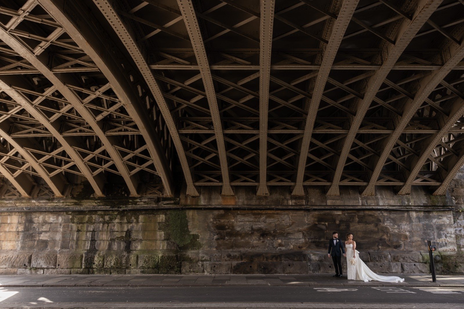 A bride and groom standing under a large bridge, with the bride in a white wedding gown and the groom in a tuxedo, on an urban street.