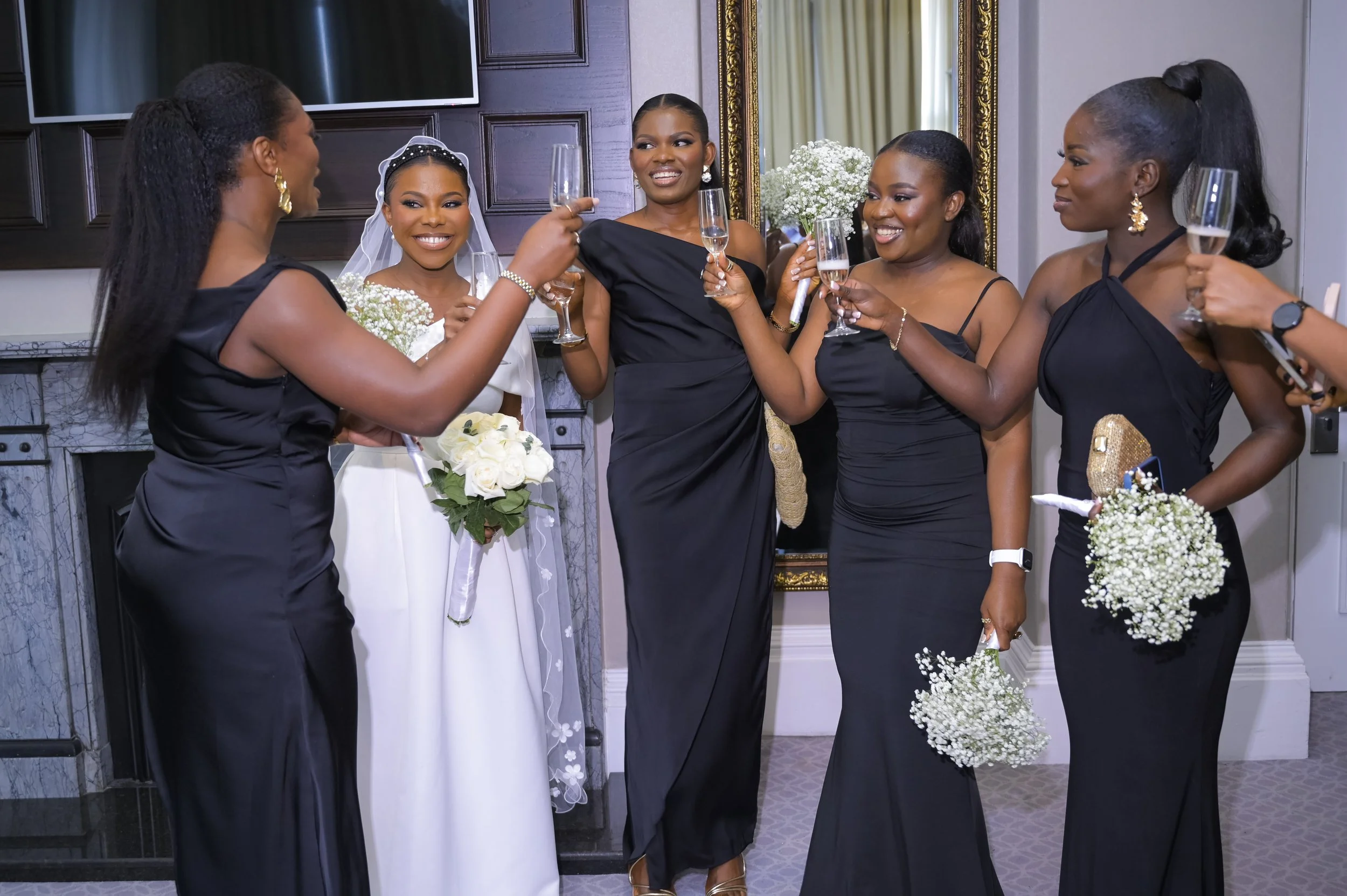 Group of women celebrating at a wedding, toasting with champagne glasses, with the bride holding a bouquet of white roses, in elegant black dresses and a white wedding gown.