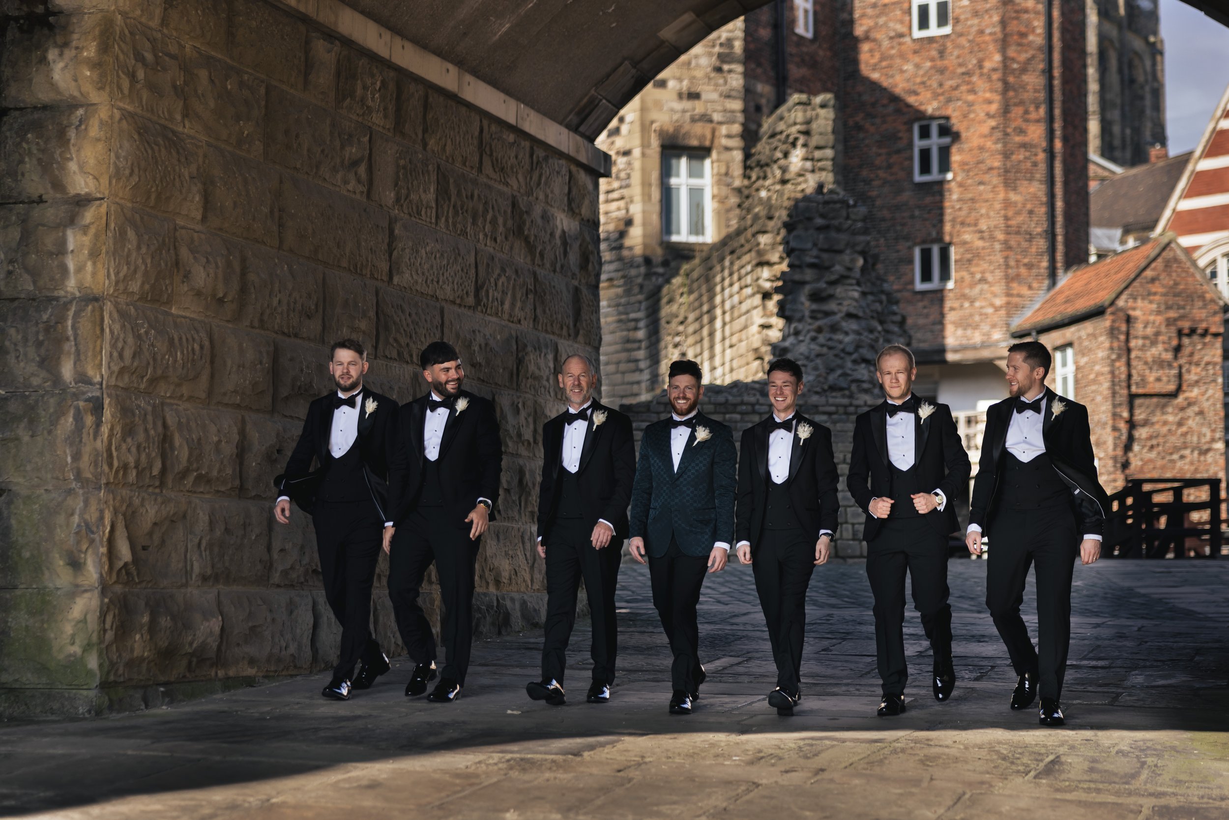 A group of seven men in tuxedos walking together outdoors near an old stone bridge and brick buildings, smiling and enjoying each other's company.