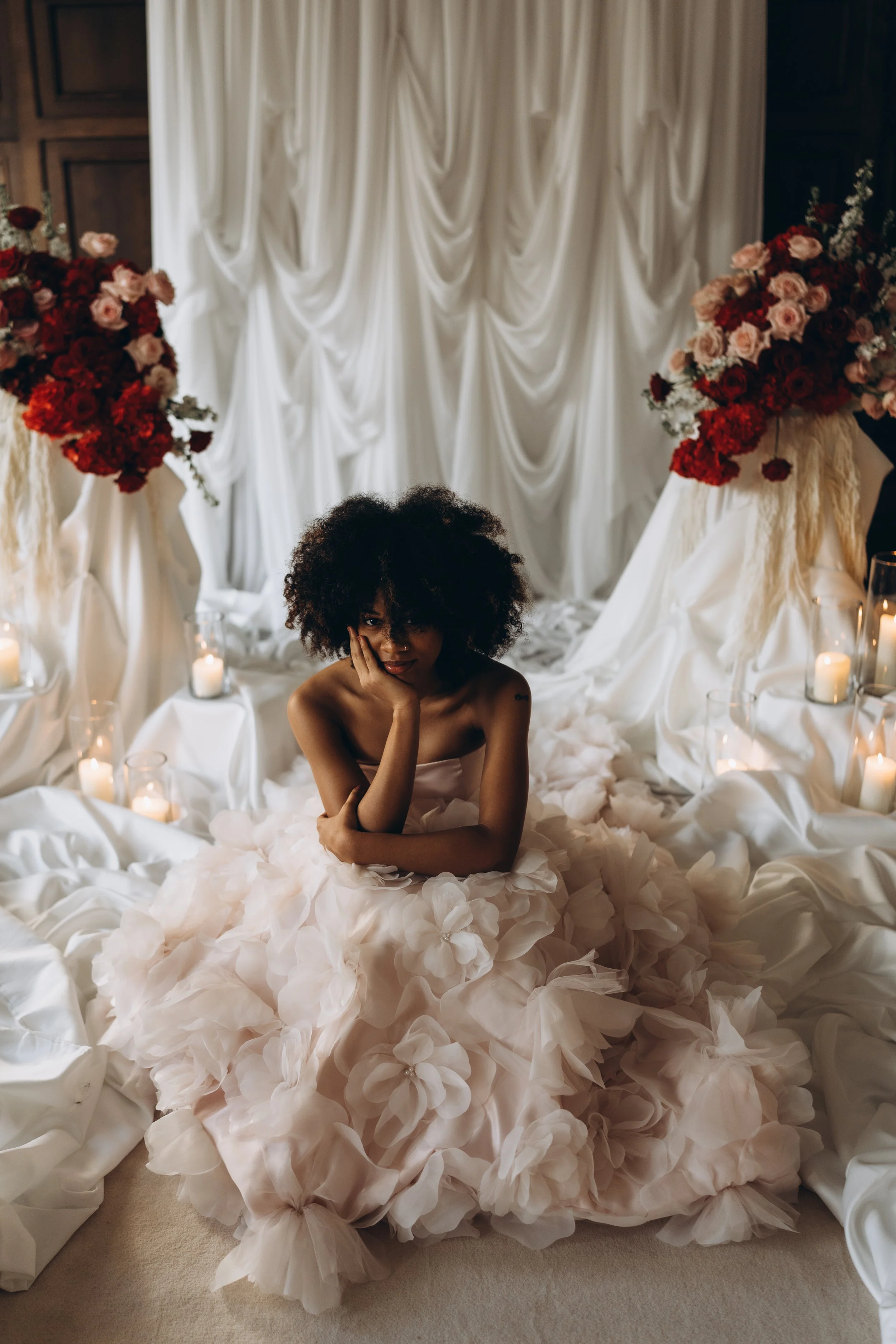 A woman with curly black hair sitting on the floor in a white wedding dress with ruffled skirt. She is in front of a white draped backdrop with large floral arrangements and candles.
