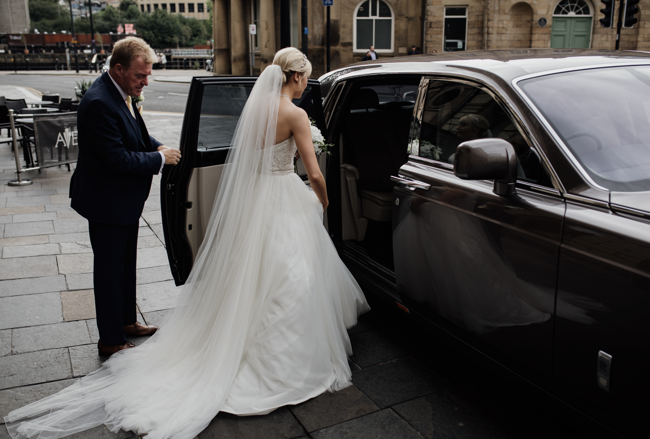 A bride with a long white wedding gown and veil getting into a black car, assisted by a man in a suit, on a city street with buildings in the background.