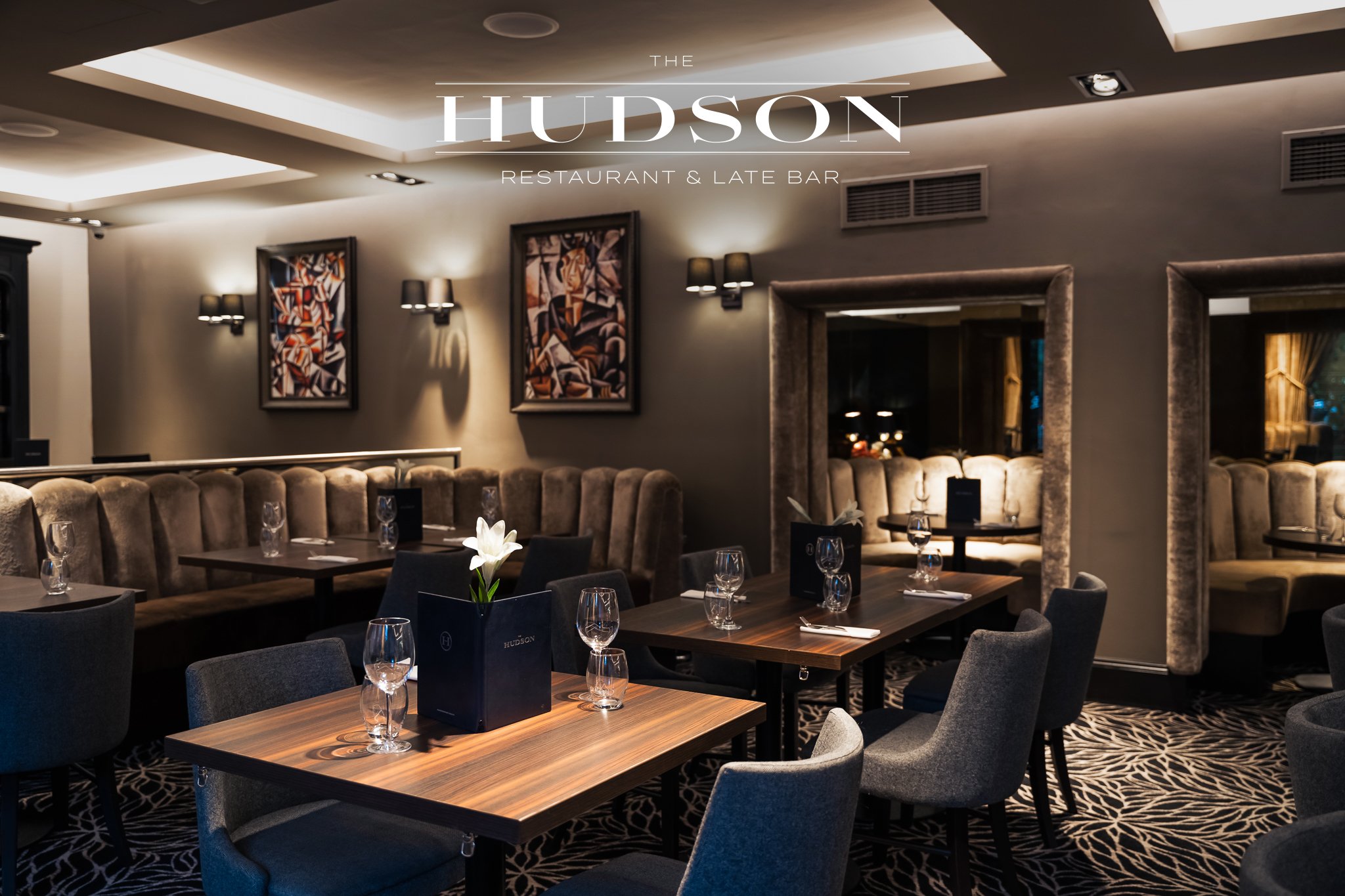 Interior of The Hudson Restaurant & Late Bar with dim lighting, artwork on gray walls, and set tables with wine glasses and menus.