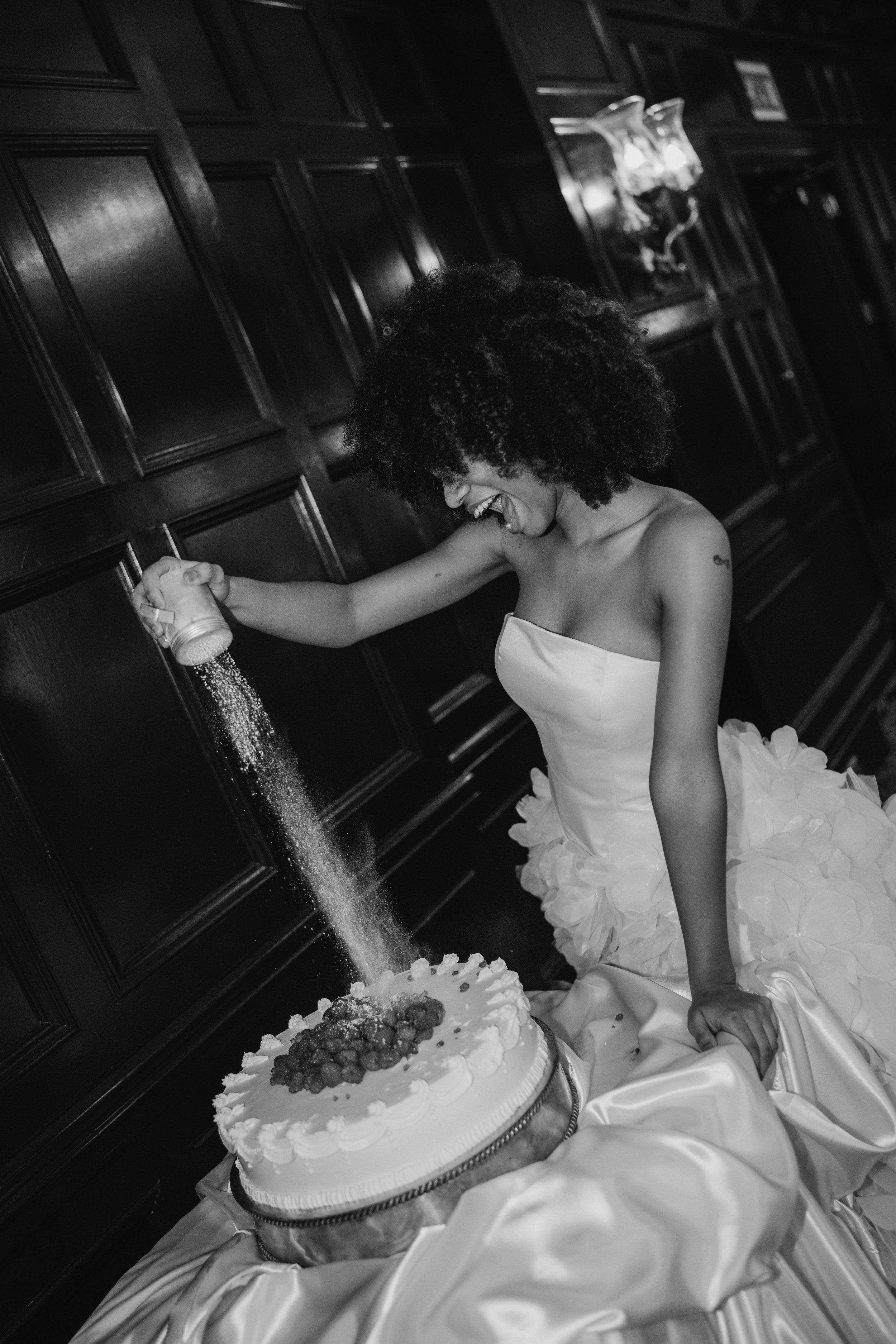 A woman in a strapless dress with a ruffled skirt is smiling happily as she sprinkles something on a cake. The scene is set indoors with dark wooden wall paneling and a decorative wall sconce in the background.