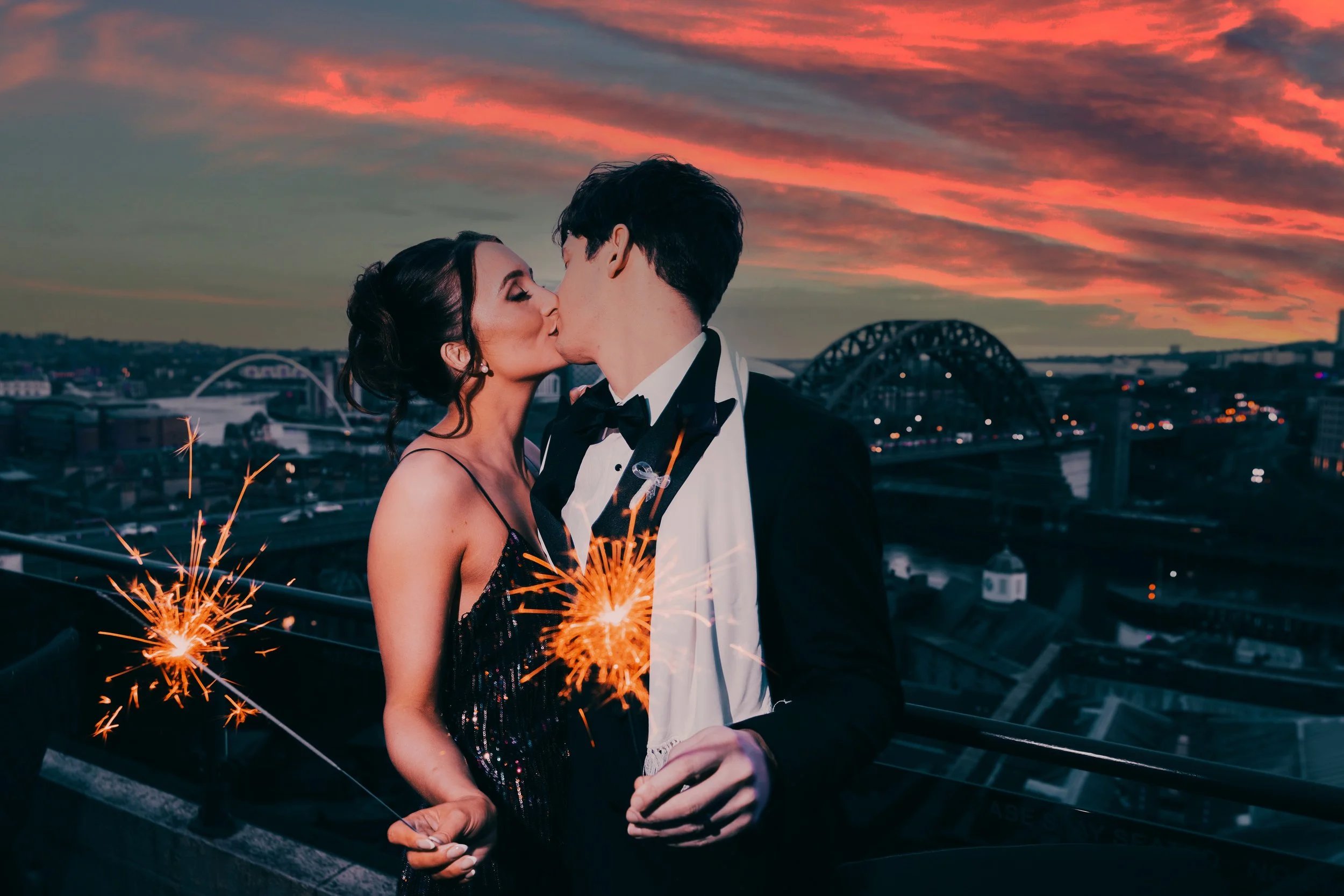 A couple dressed in formal attire, sharing a kiss on a rooftop at sunset, holding sparklers with an illuminated cityscape and bridge in the background.