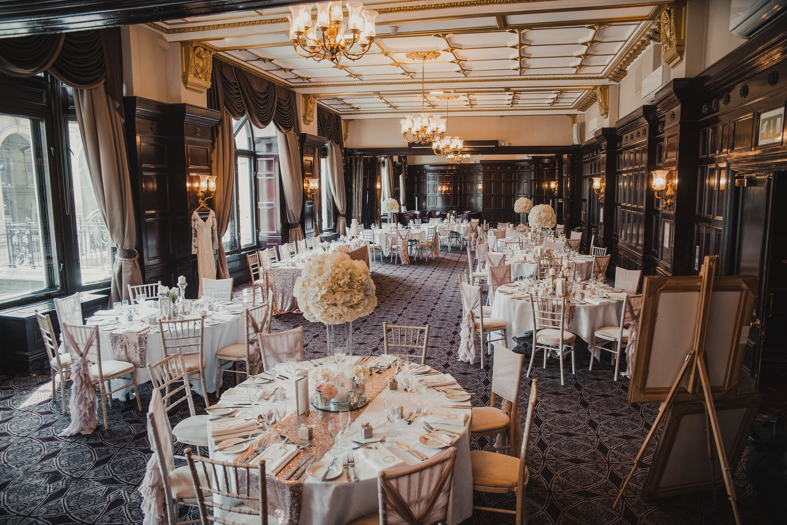 Elegant banquet hall set up for a wedding reception with round tables decorated with white tablecloths, floral centerpieces, and place settings, featuring dark wood-paneled walls, tall windows with curtains, and chandeliers hanging from the ceiling.