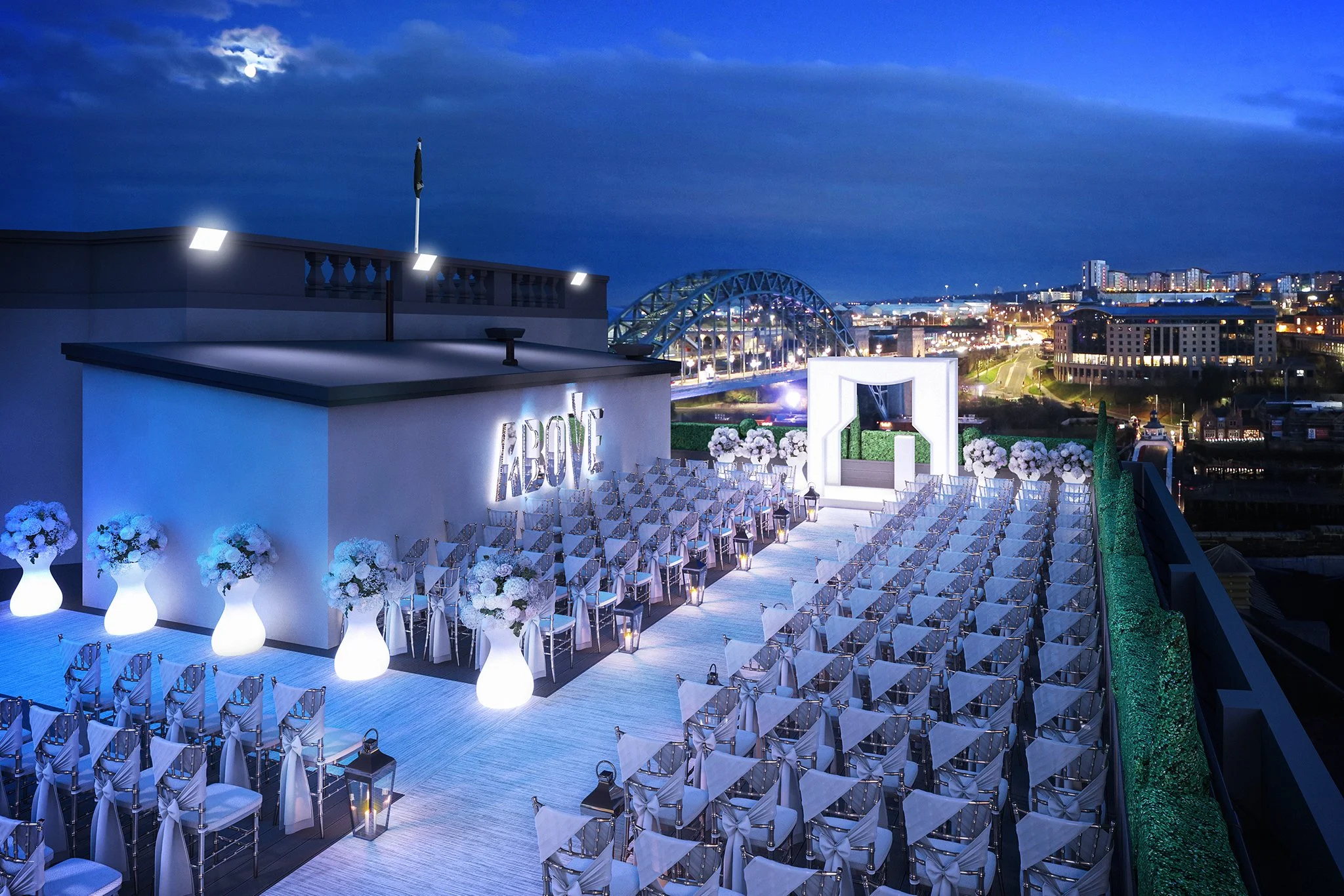Outdoor wedding setup in the evening with chairs arranged in rows, decorated with white bows, facing an illuminated arch on a balcony overlooking a cityscape with bridges and buildings under a cloudy sky with the moon visible.