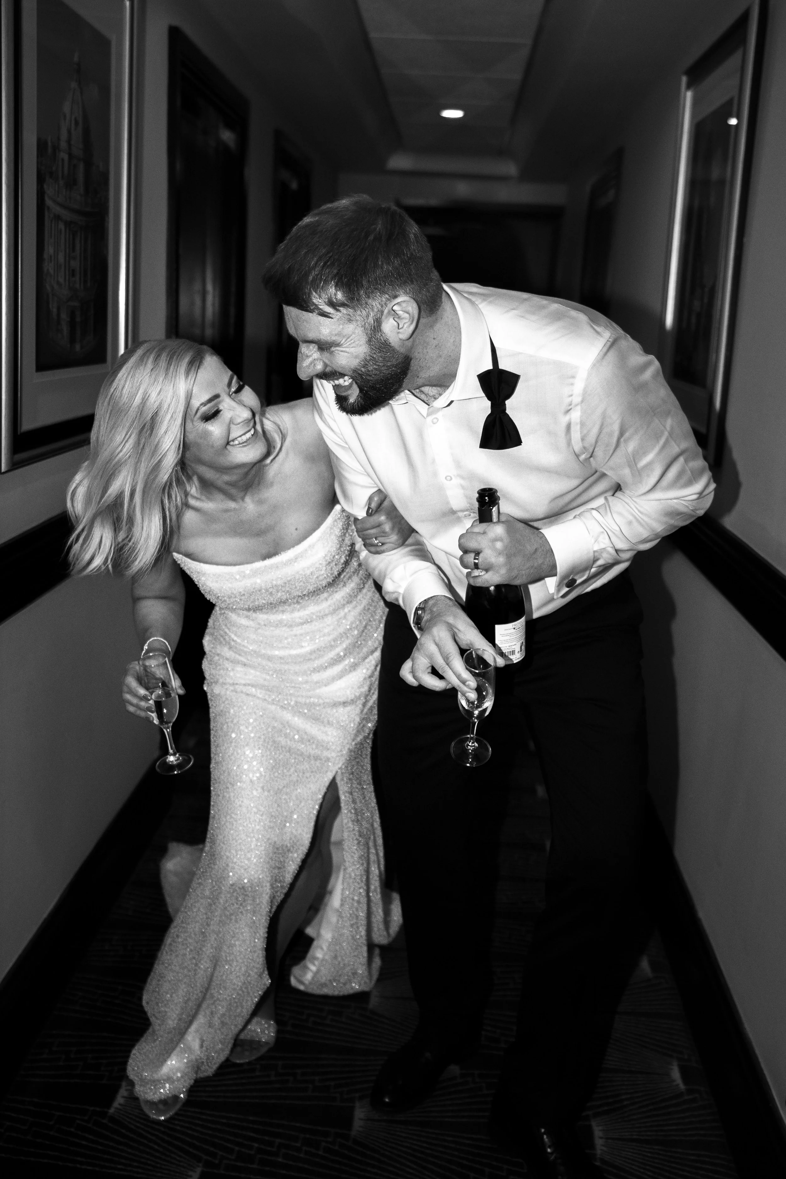 A black and white photograph of a joyful woman in a sparkling dress and a man in formal attire with a bow tie, both holding champagne glasses and laughing as they dance closely together in a corridor with framed pictures on the walls.