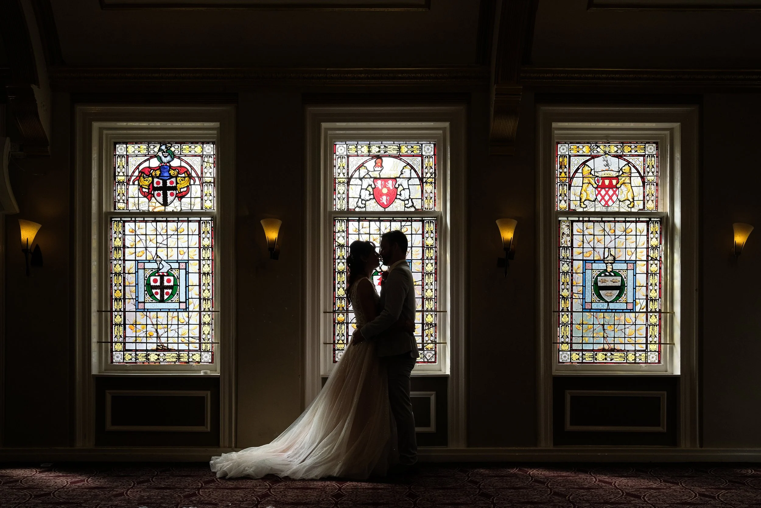 Silhouette of a bride and groom standing close together inside a room with large stained-glass windows depicting coats of arms, natural light streaming through, creating a backlit scene.