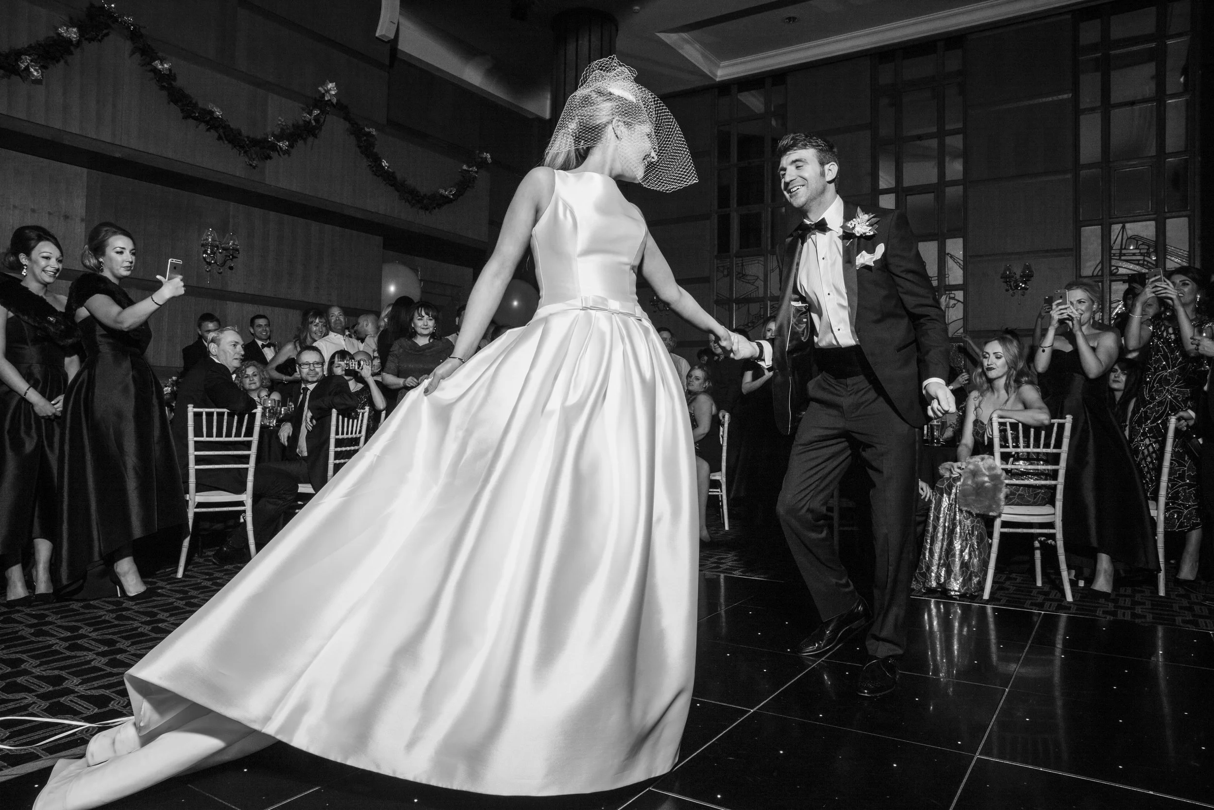 A bride and groom dancing at their wedding reception, surrounded by guests in a decorated hall.