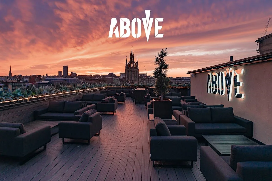 Rooftop lounge during sunset with black outdoor sofas, potted trees, and illuminated 'ABOVE' sign, overlooking city skyline with a church and sunset sky.
