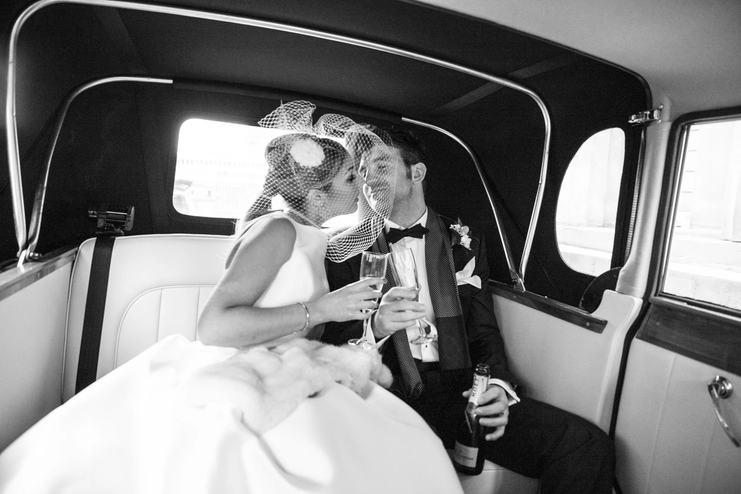 A bride and groom sitting inside a taxi. The bride is holding a champagne flute and is leaning in to kiss the groom, who is smiling. Car's interior and window are visible.