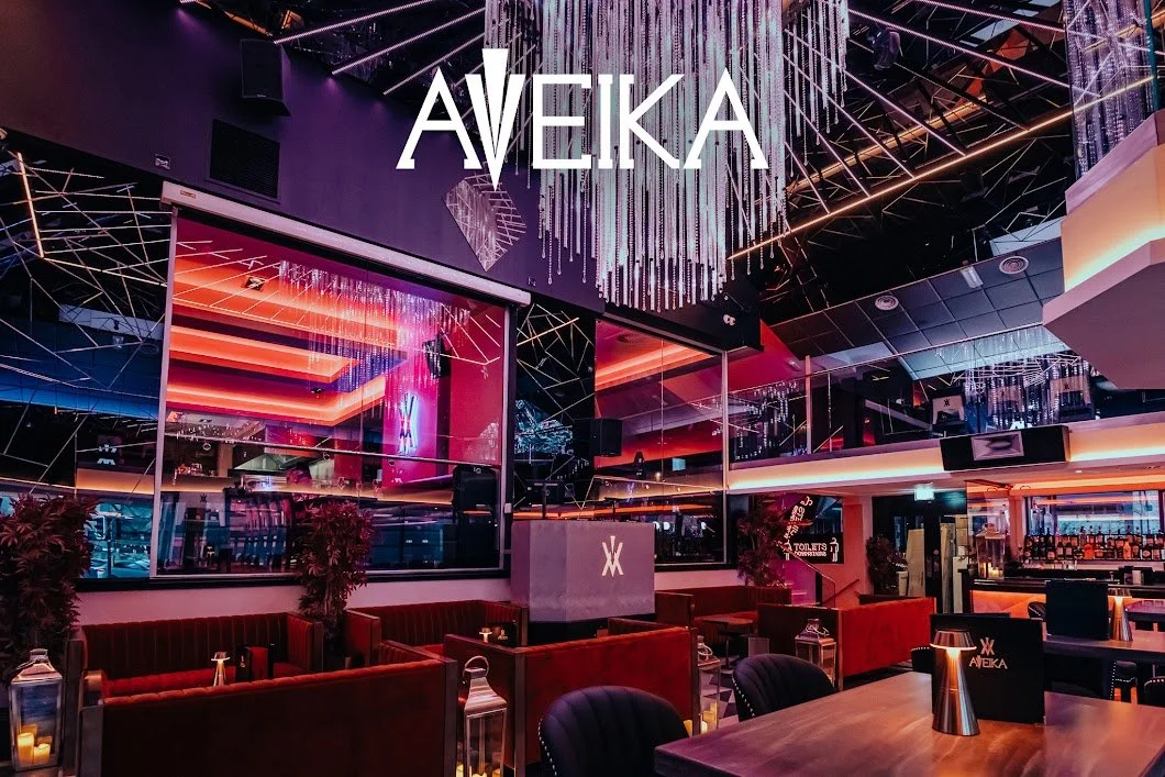Interior of a modern nightclub or lounge with vibrant lighting, red cushioned seating, and mirrors, featuring the name Aveika prominently displayed.