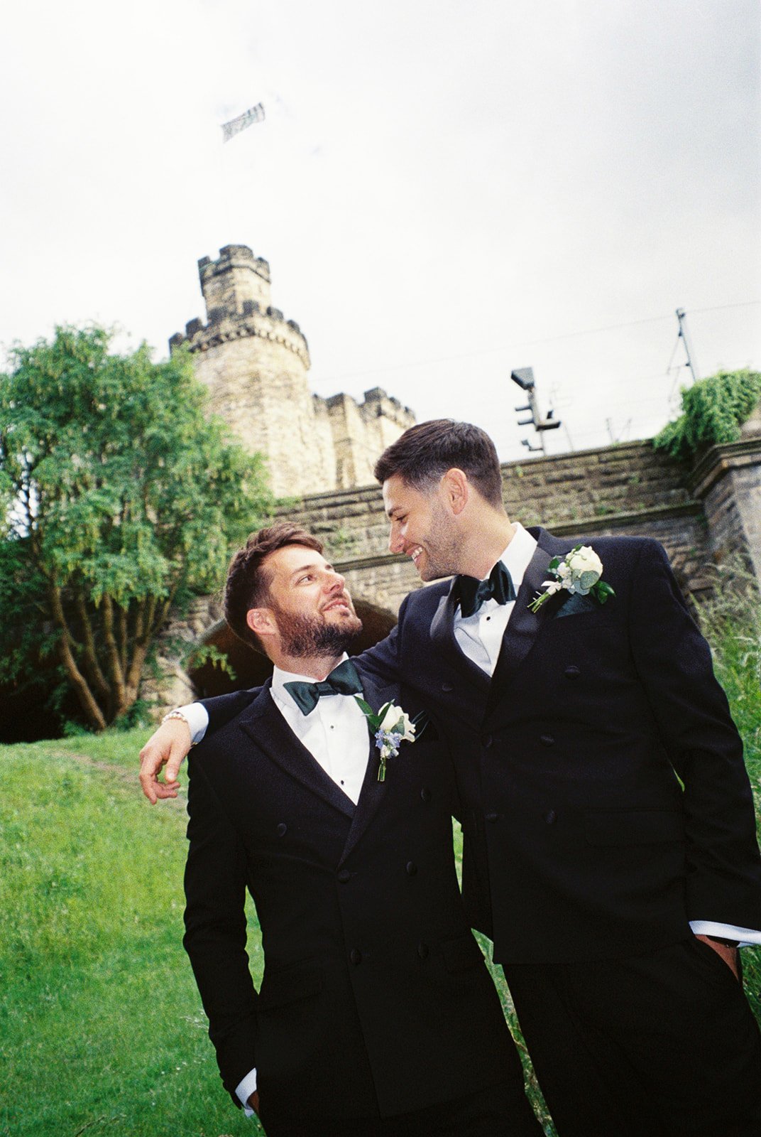 Two men in tuxedos with boutonnieres, smiling and embracing each other outdoors in front of a stone castle.