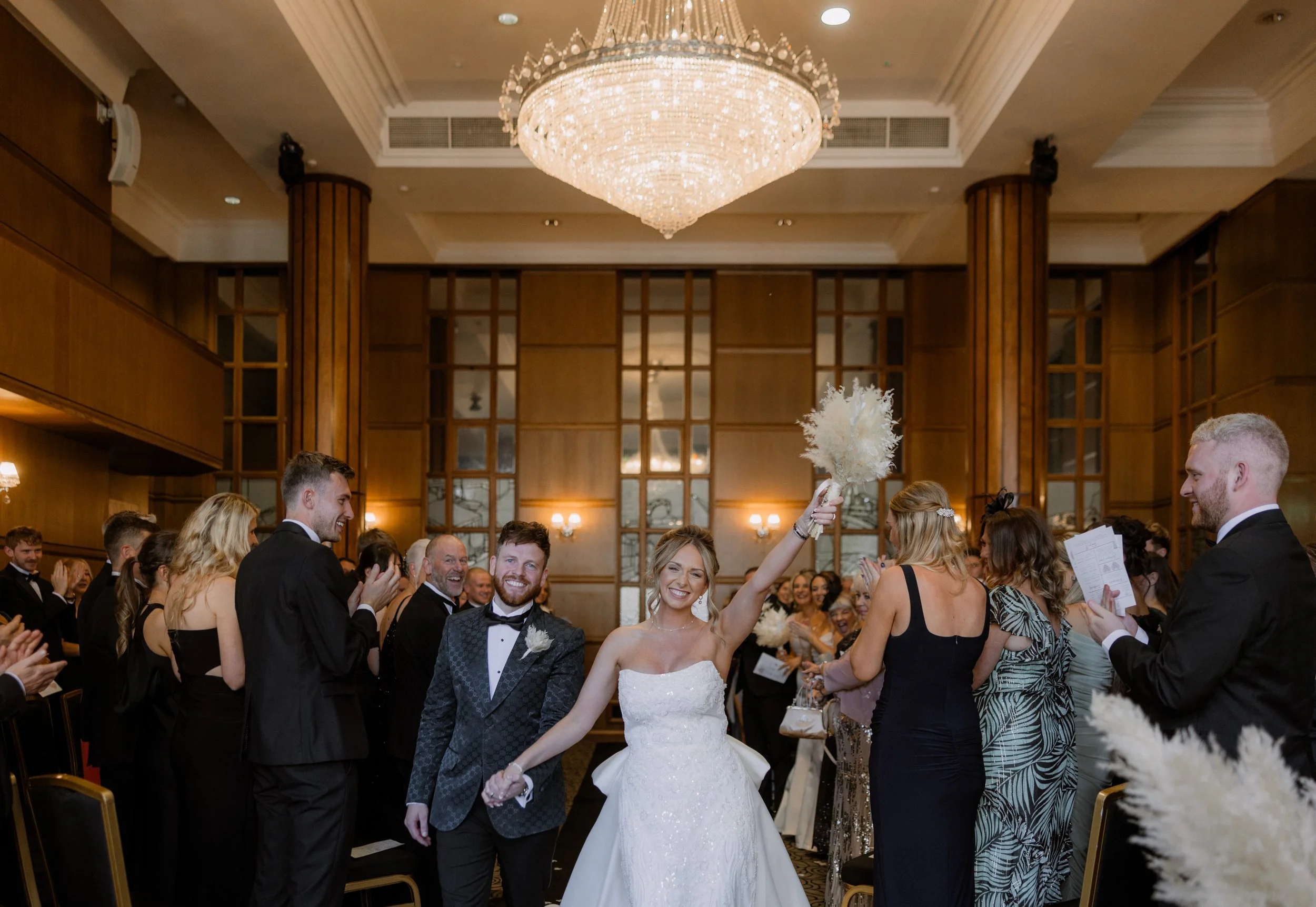 A bride in a white wedding gown and a groom in a tuxedo smiling and holding hands at a wedding reception, surrounded by guests in formal attire, inside an elegant hall with a large chandelier overhead.