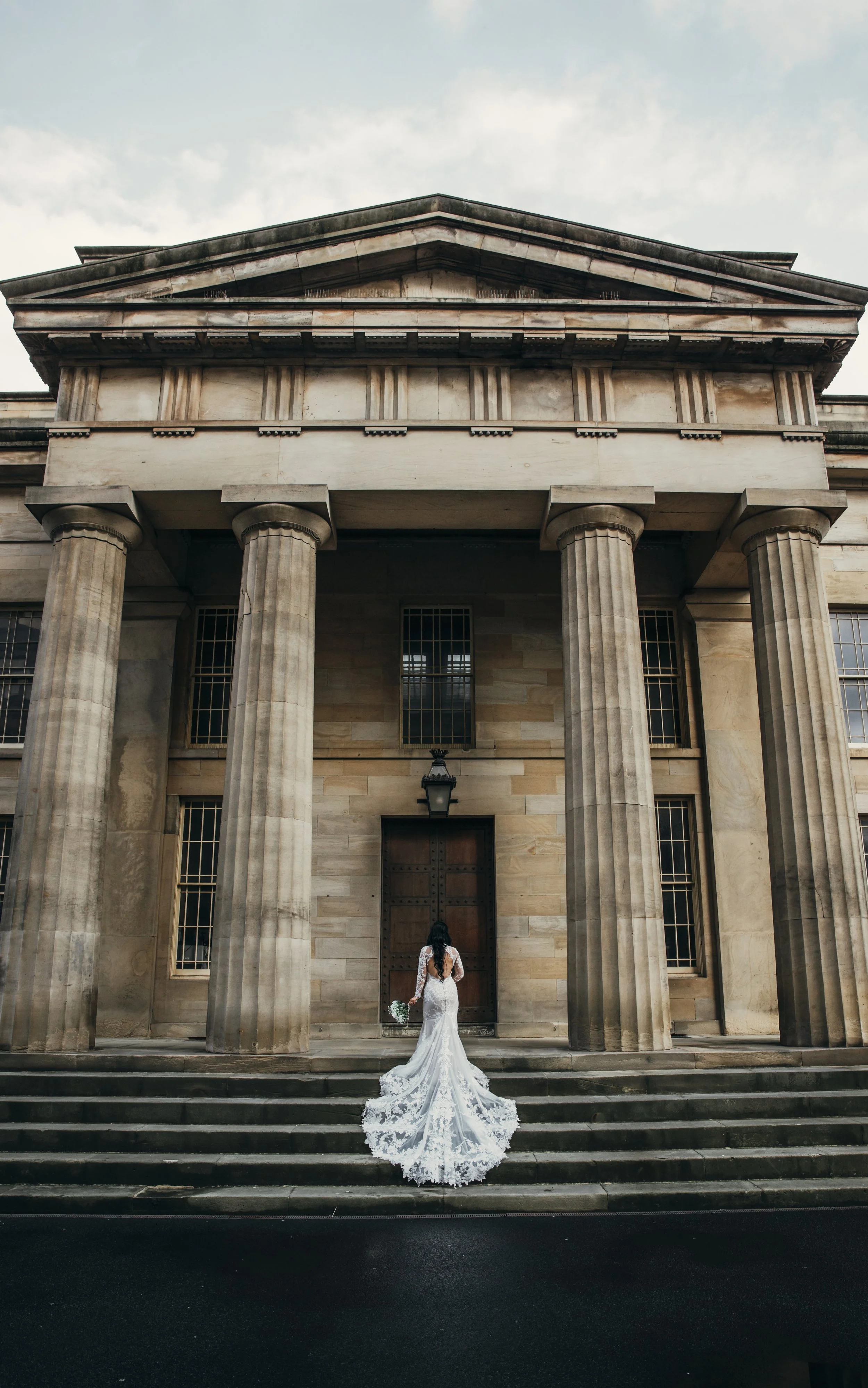 A bride in a white wedding dress holding a bouquet stands at the entrance of a large, historic stone building with tall columns and a big wooden door, under a cloudy sky.