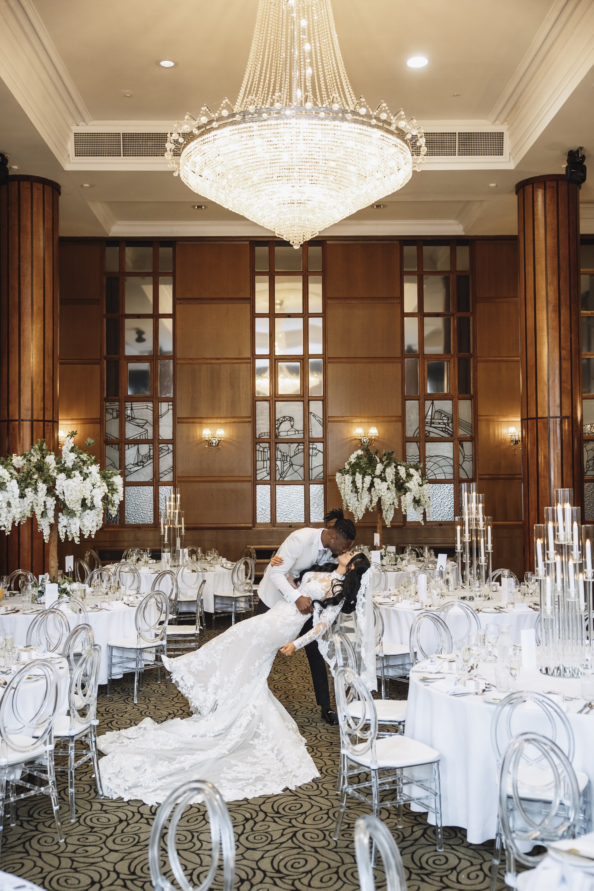 A bride and groom are dancing in a luxurious banquet hall with wooden accents, a large chandelier, and floral centerpieces.
