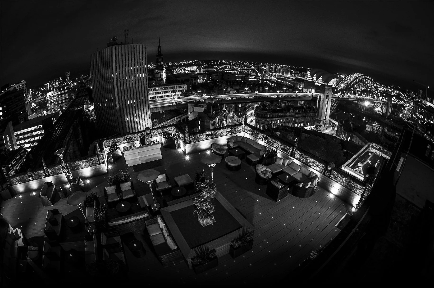 Nighttime cityscape view from a rooftop lounge, with outdoor seating, illuminated planters, and city lights in the background.