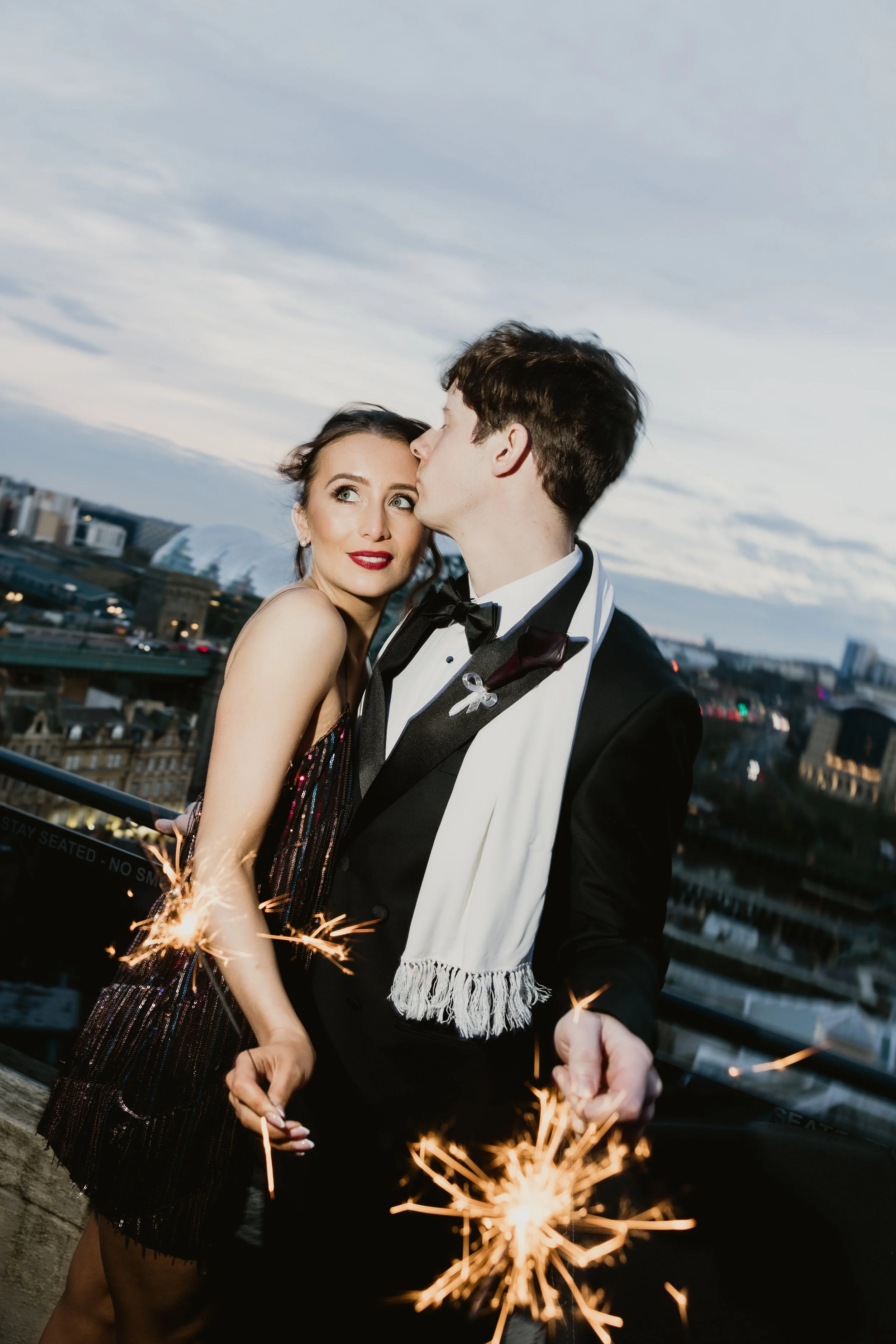 A man and woman dressed in formal attire celebrating on a rooftop at dusk, holding sparklers with a city skyline in the background.