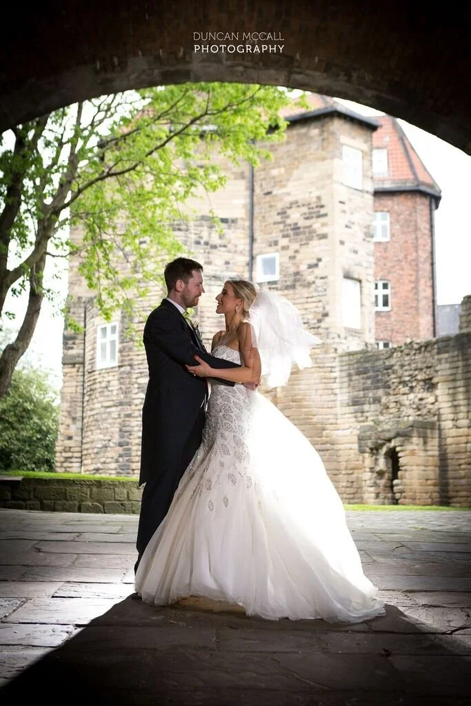 A bride and groom stand under a stone archway, embracing and looking at each other, with an old stone building and trees in the background.