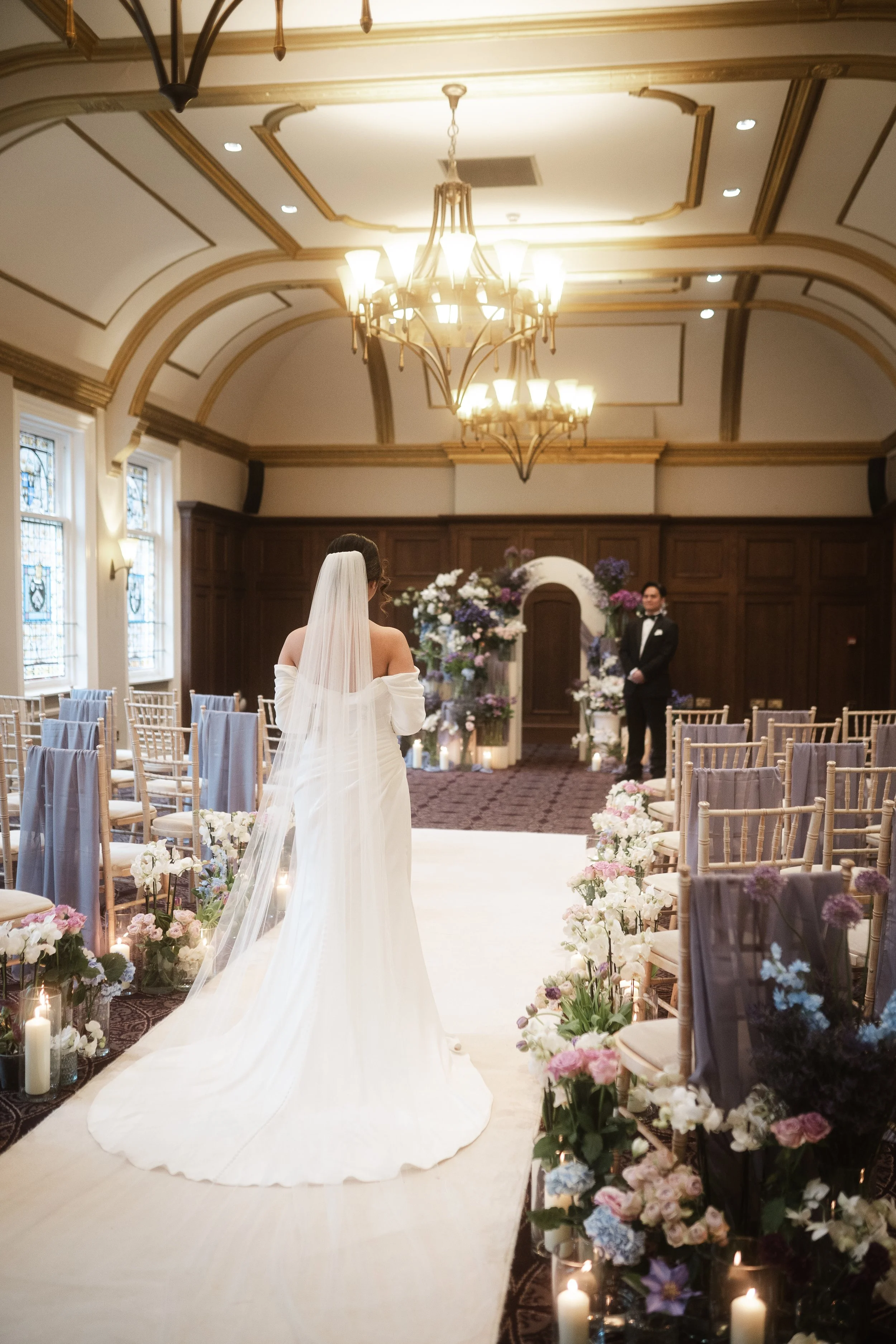 A bride in a white wedding dress and veil walking down the aisle in a decorated wedding venue, with chairs on either side decorated with flowers and candles, and a man in a tuxedo standing in the background.