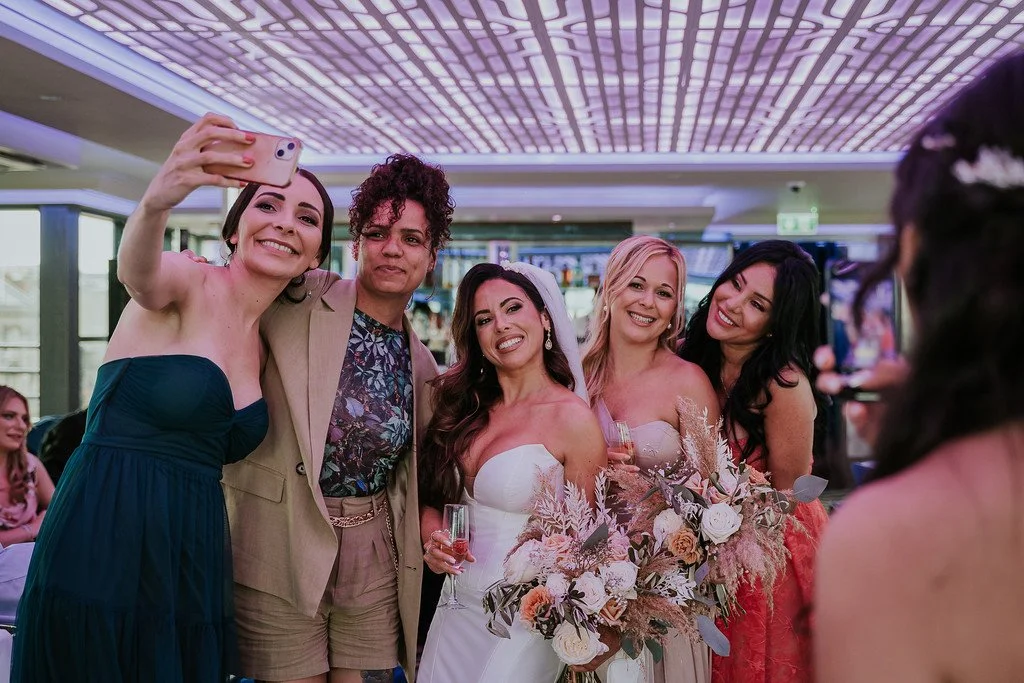 Five women at a wedding, taking a selfie with a bride holding a bouquet of flowers in the center. The women are smiling and dressed in formal attire.