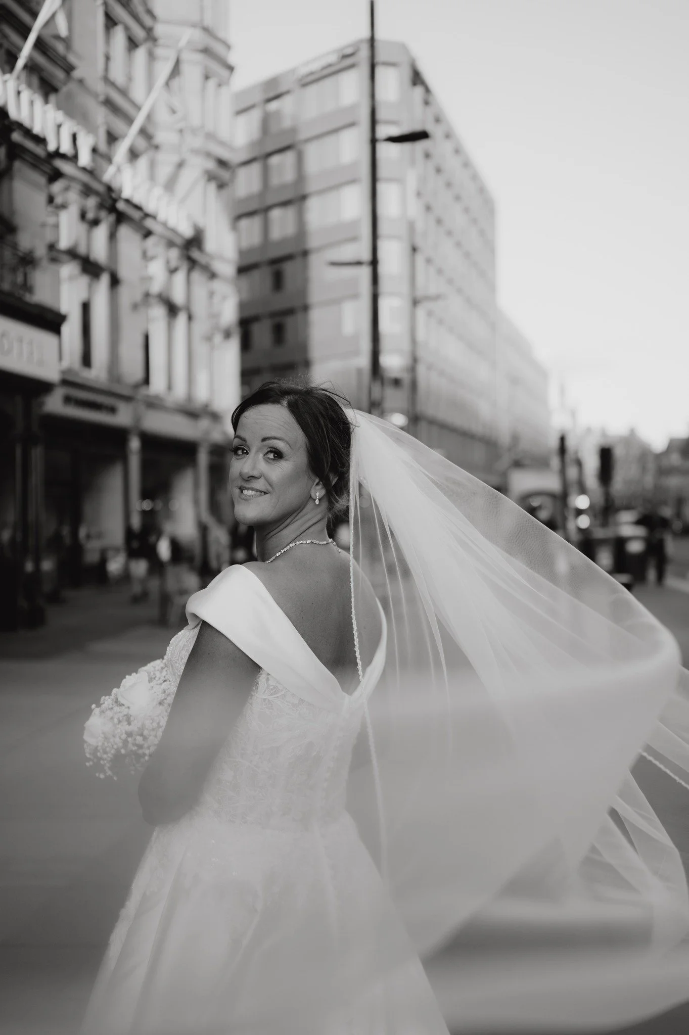 A smiling bride in a wedding dress and veil standing on a city street with buildings in the background, captured in black and white.