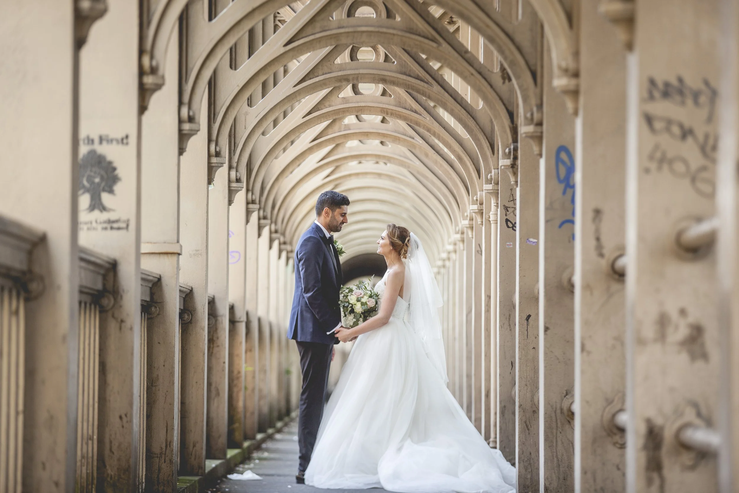 A bride and groom standing face-to-face under a series of arched stone structures, holding hands, surrounded by graffiti on the walls.