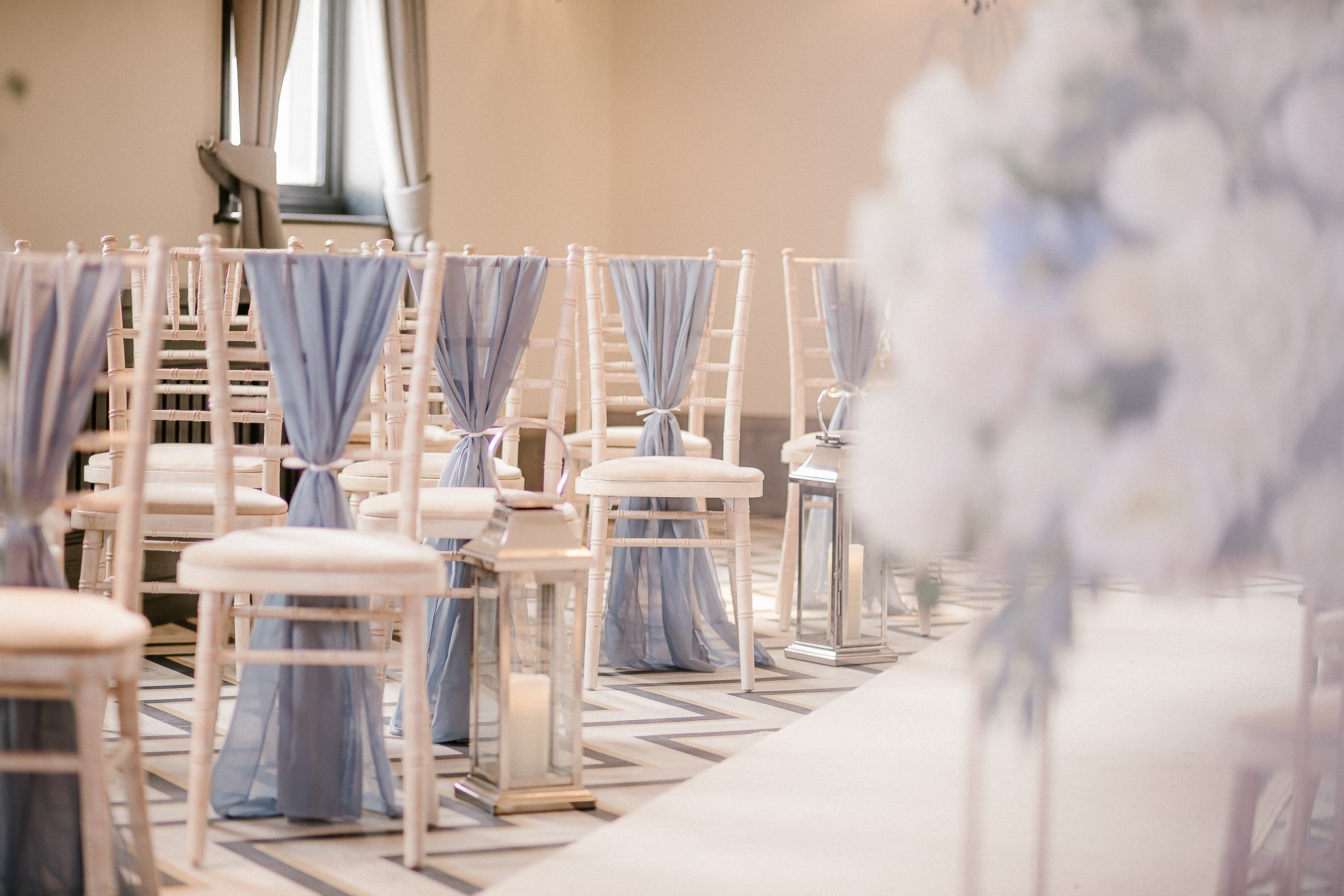 Rows of white chairs with blue drapes tied to the back at a decorated event venue with a beige wall and a window with curtains.