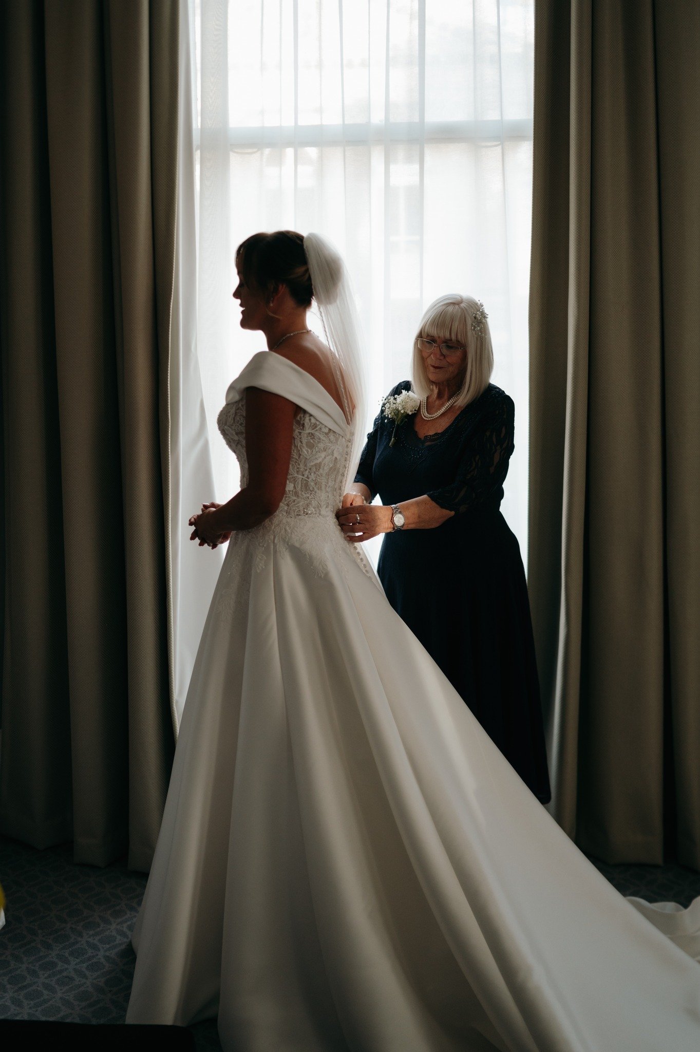 A bride in a wedding dress with a veil is being helped by an older woman in a black dress and glasses, standing in front of a window with sheer curtains.