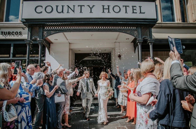 Bride and groom leave County Hotel under a shower of confetti as guests celebrate.
