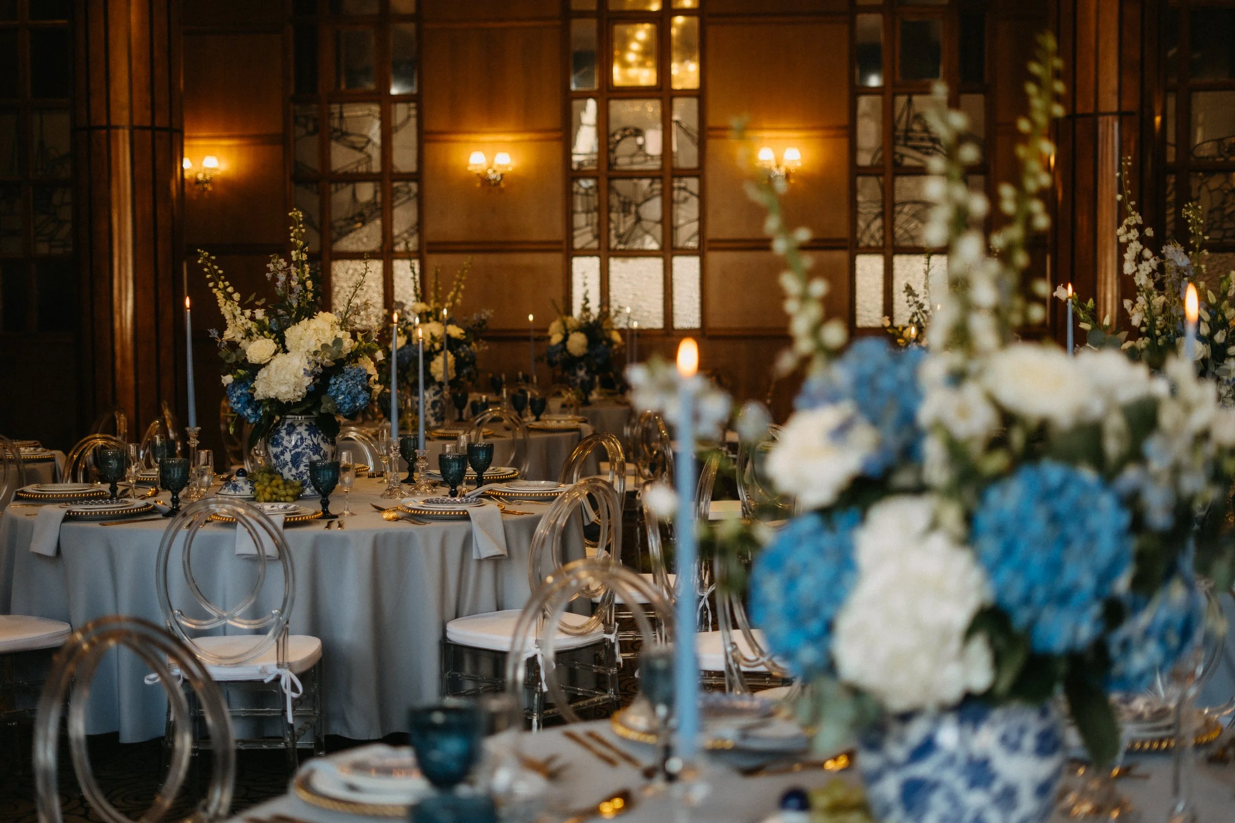 Elegant indoor banquet setup with round tables decorated with tall floral centerpieces of white and blue flowers, gold-rimmed plates, and blue drinking glasses, set against a wooden paneled wall with large mirrors and warm wall sconces.