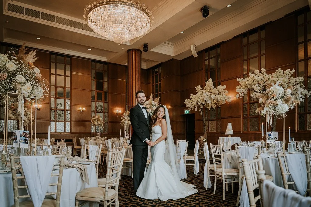 Bride and groom holding hands and smiling at a wedding reception in a decorated banquet hall with tall floral arrangements and a chandelier.