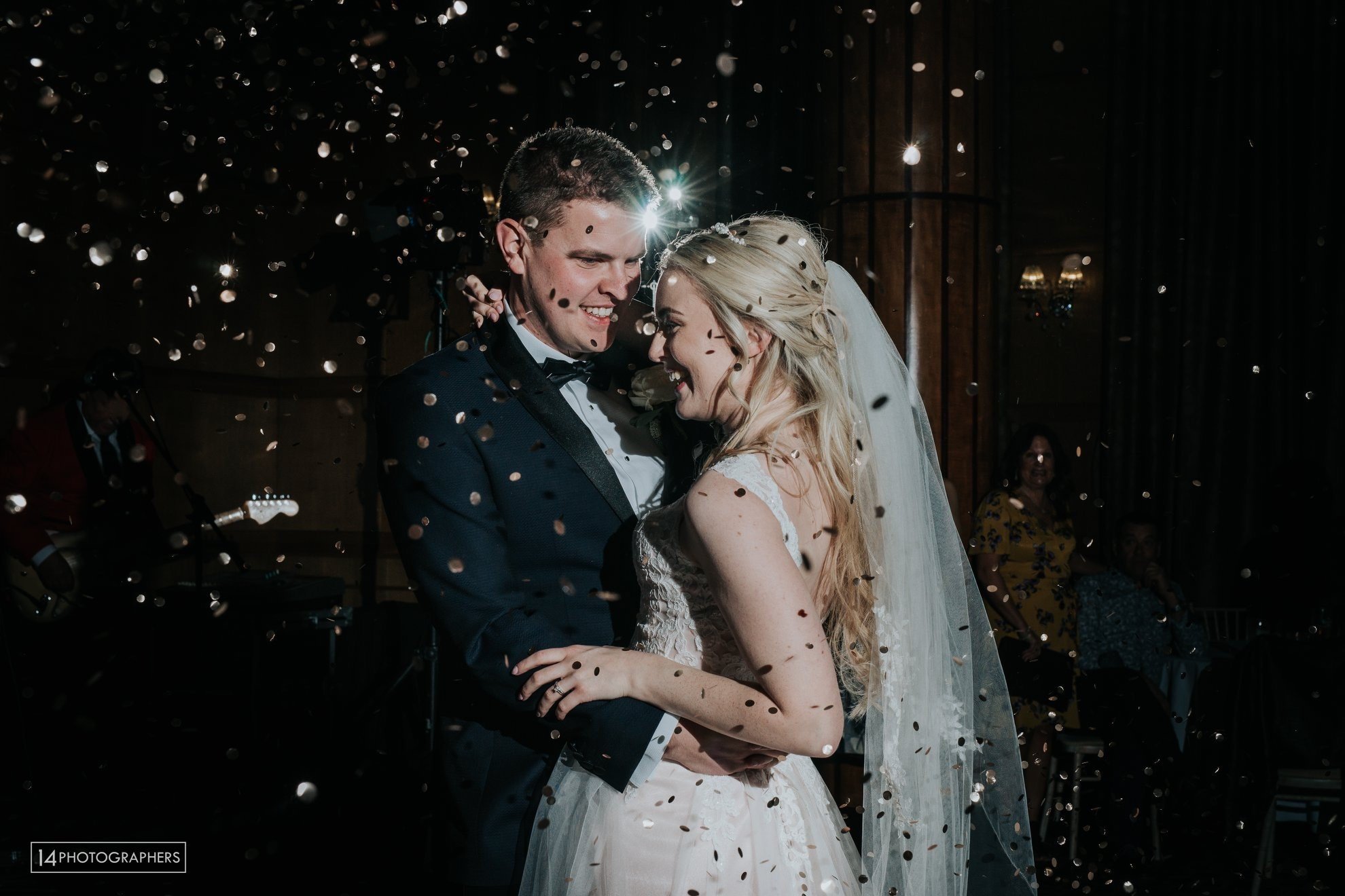 A bride and groom dancing at their wedding reception surrounded by falling confetti, with guests in the background.