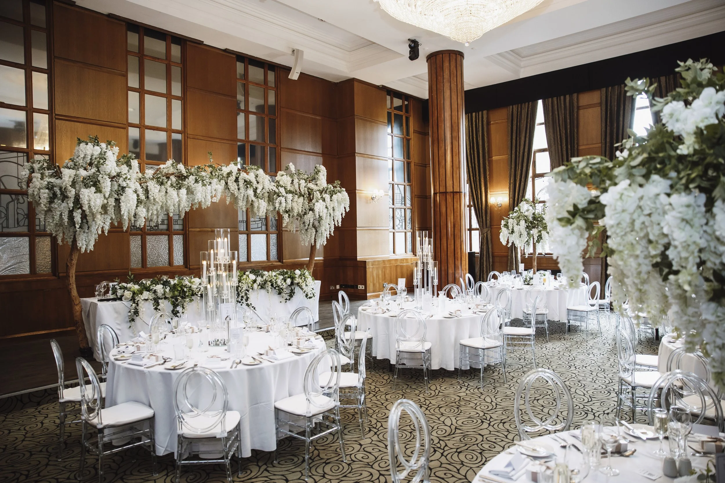 Elegant banquet hall decorated for a wedding or celebration, with round tables draped in white cloth, transparent chairs, floral centerpieces with white flowers and green foliage, tall candles, large windows with dark curtains, wooden wall paneling, 